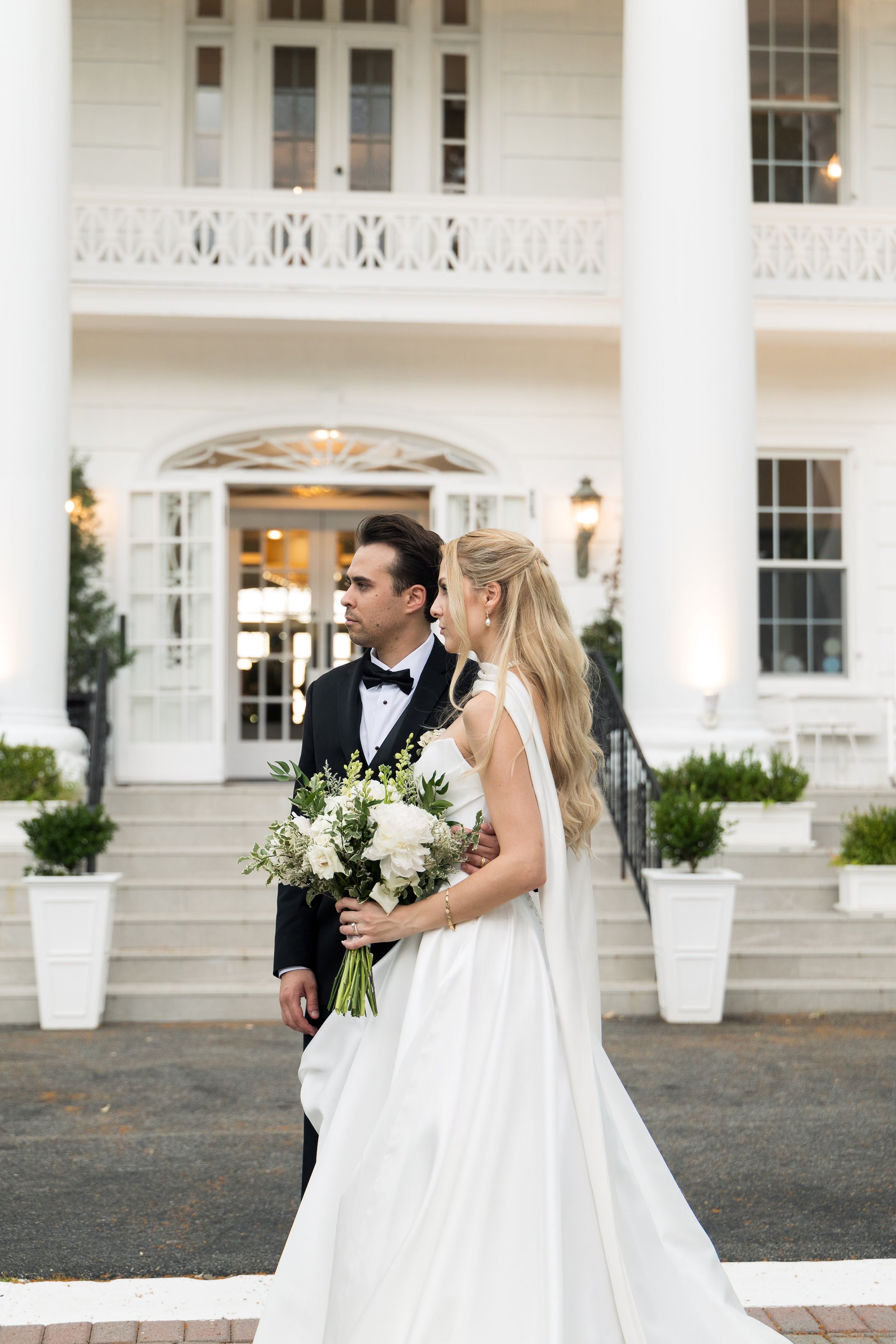 Bride and groom standing in front of a white building with columns, surrounded by potted plants, a staircase, and large windows, during a wedding ceremony.
