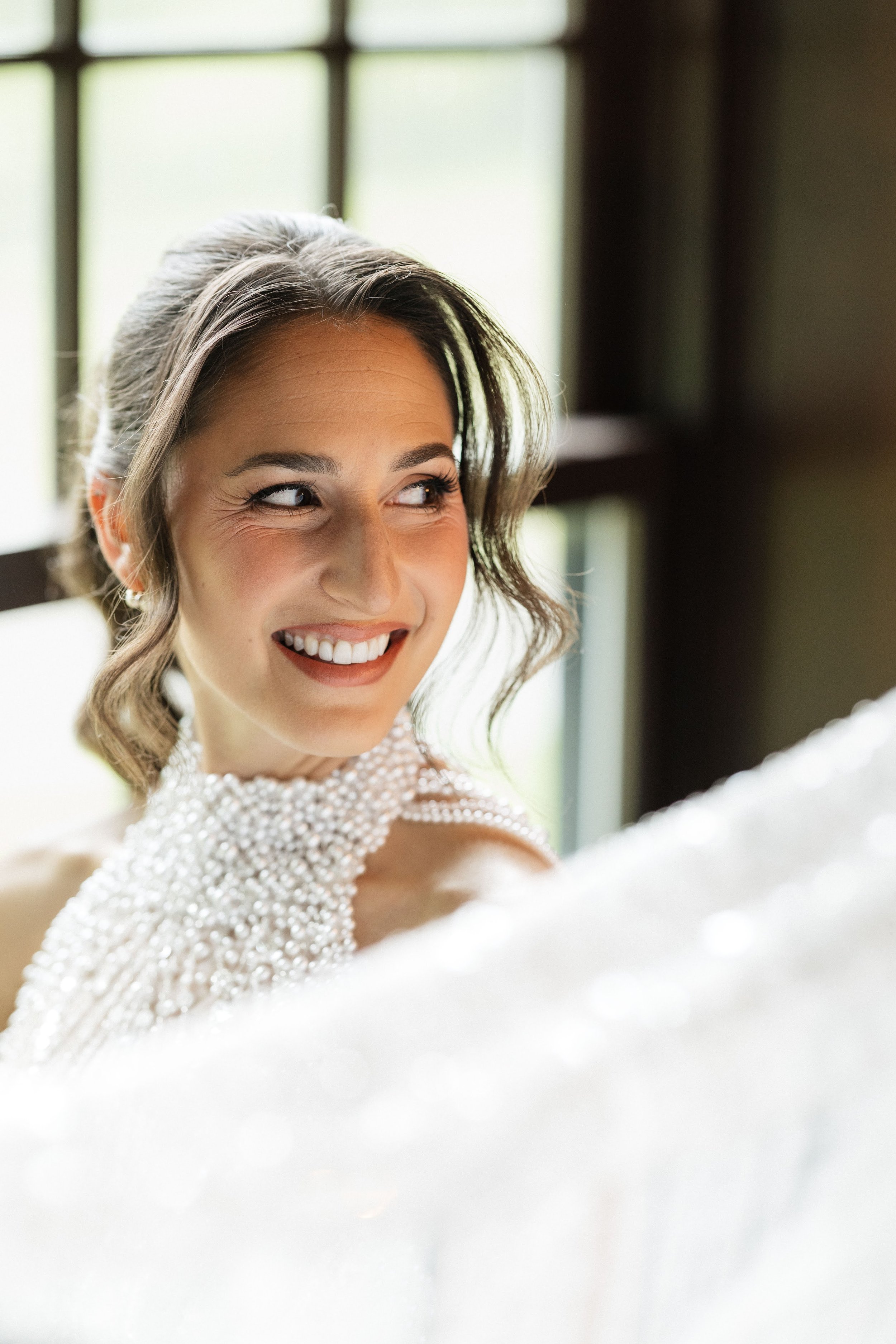 A smiling woman with short, wavy hair looking to her left, wearing a beaded or pearl dress, standing near a window with green scenery outside.