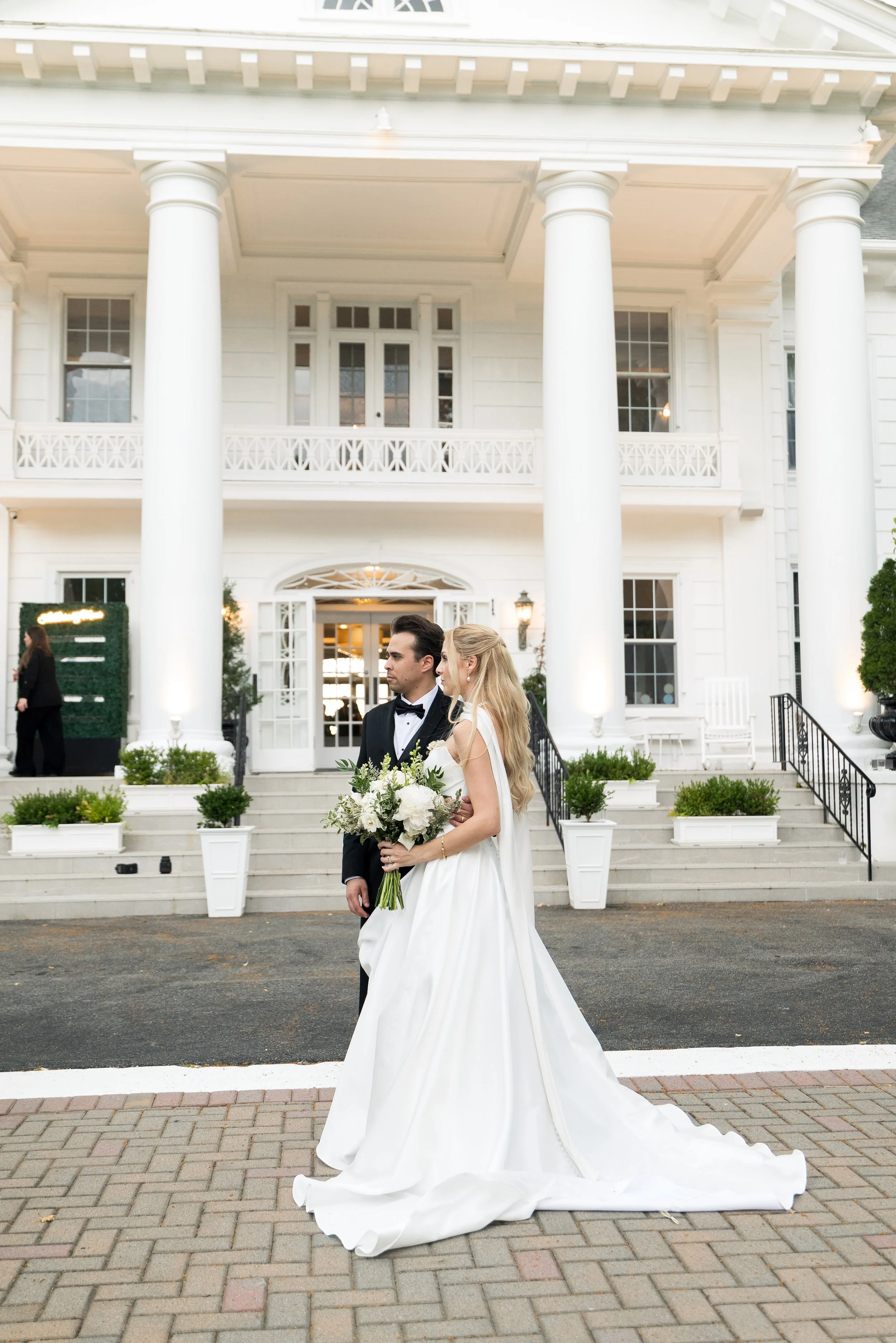 A bride and groom standing in front of a white, two-story house with large columns and a porch, during a wedding ceremony. The bride is holding a bouquet of white flowers, and the groom is dressed in a tuxedo. The house has multiple windows and decor