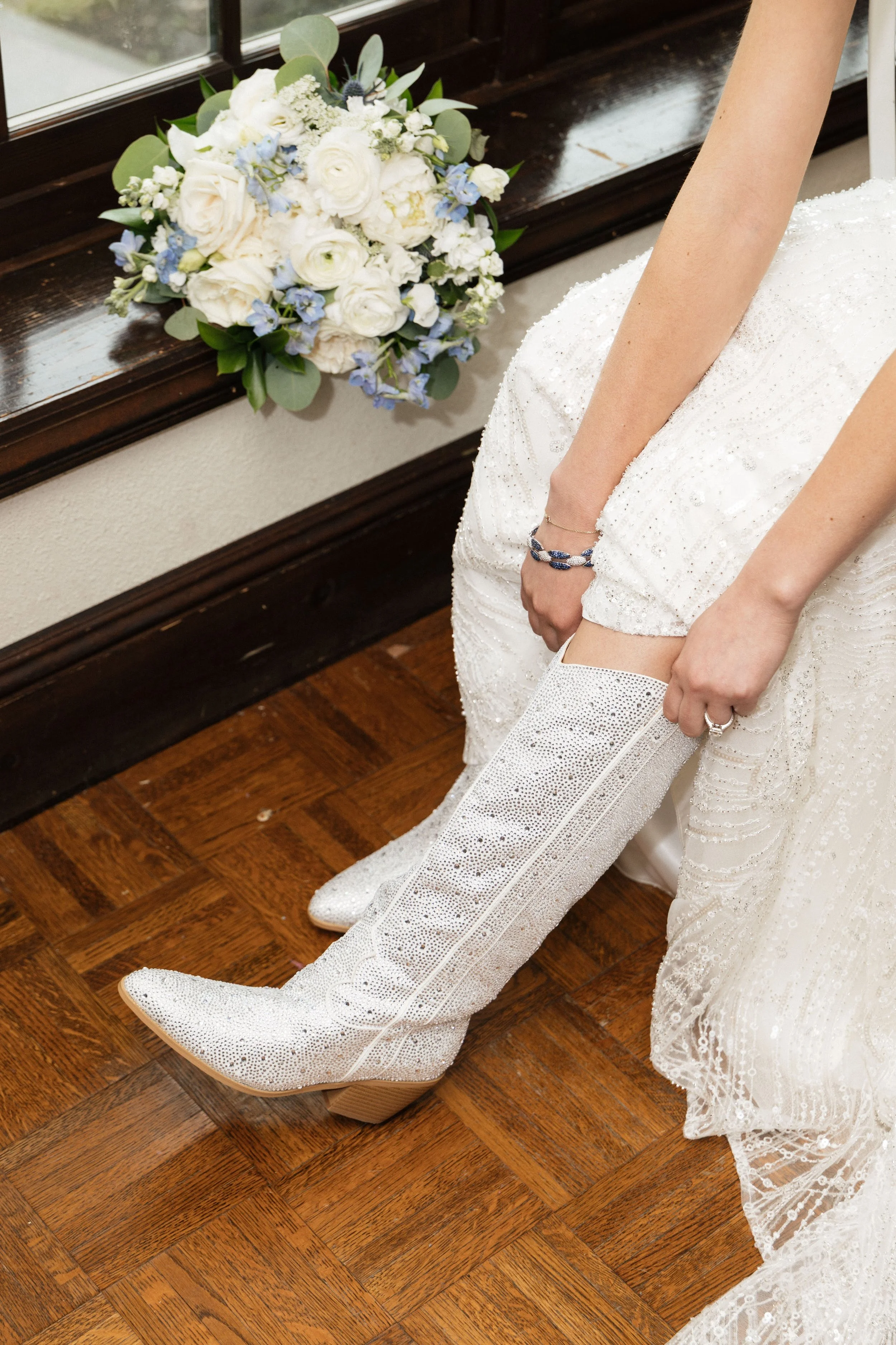 A bride wearing a sequined white wedding dress and white cowboy boots with a pointed toe, sitting on a wooden floor next to a bouquet of white and blue flowers near a window.