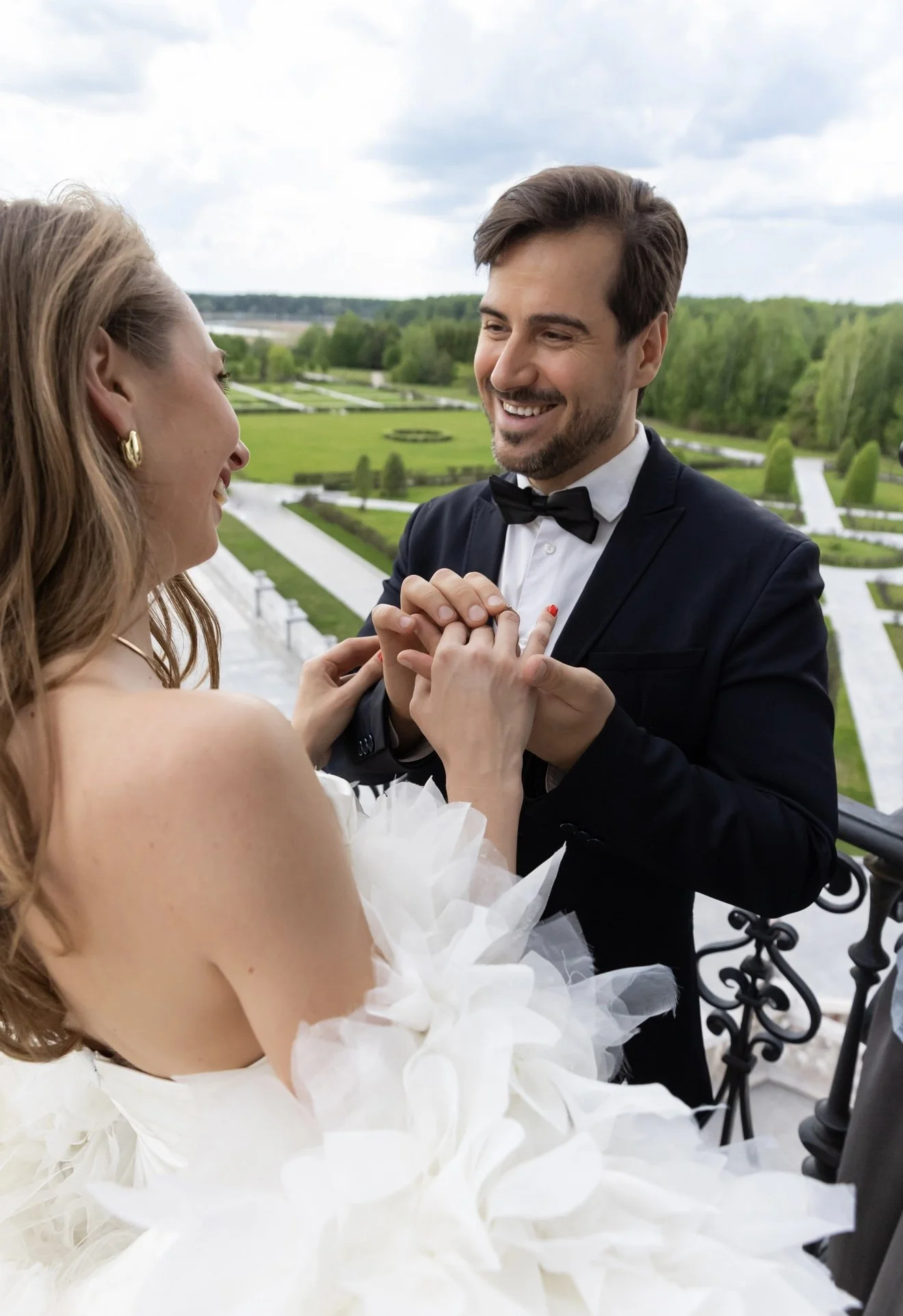 A man and woman dancing outdoors on a balcony with a lush green garden in the background. The woman is wearing a white dress with puffed sleeves, and the man is in a tuxedo with a bow tie. They are smiling and holding hands.