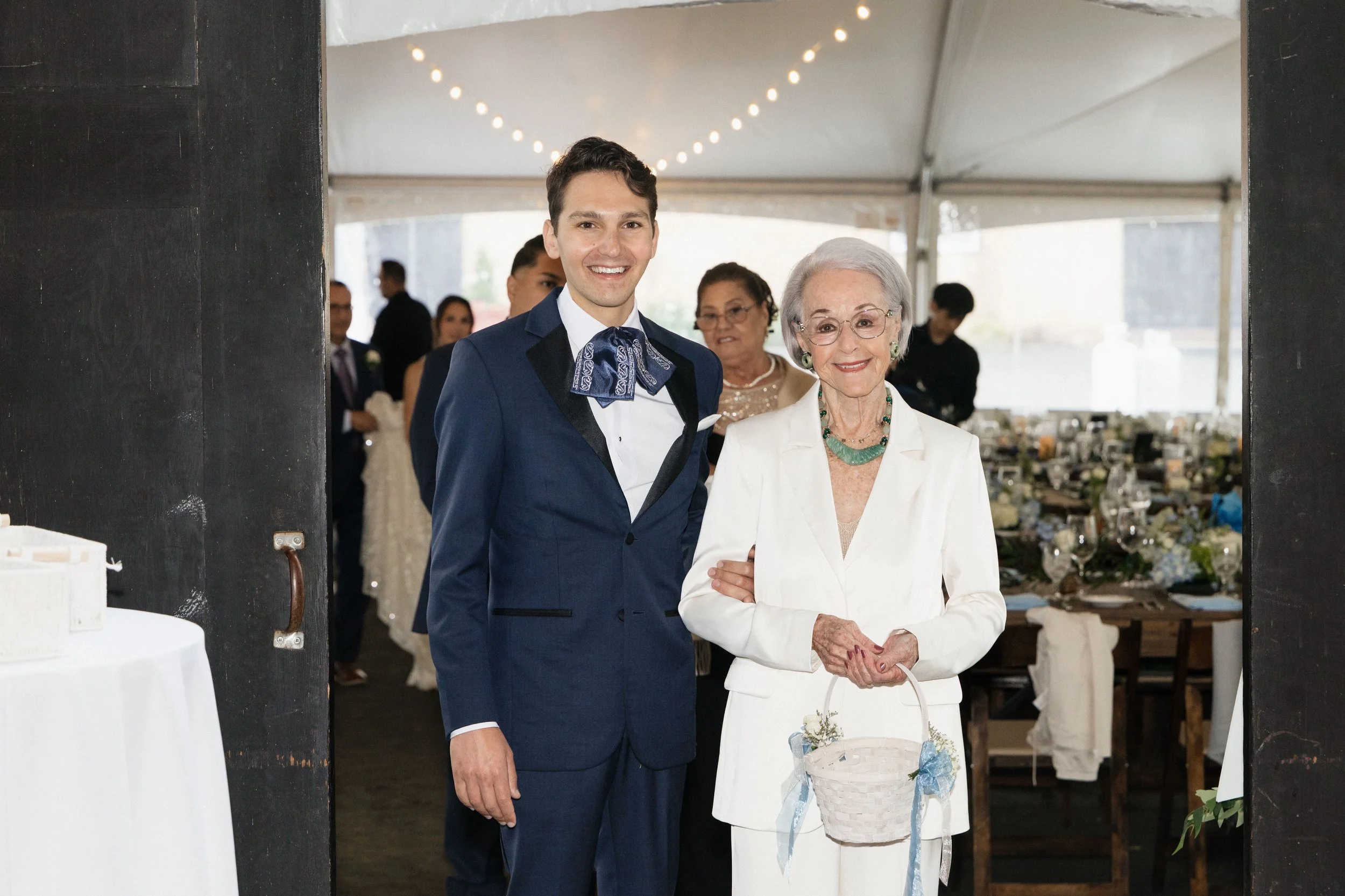 A young man in a navy suit and blue patterned bowtie standing beside an elderly woman in white attire, both smiling at a celebration event inside a large tent decorated with string lights. There are other guests and tables in the background.