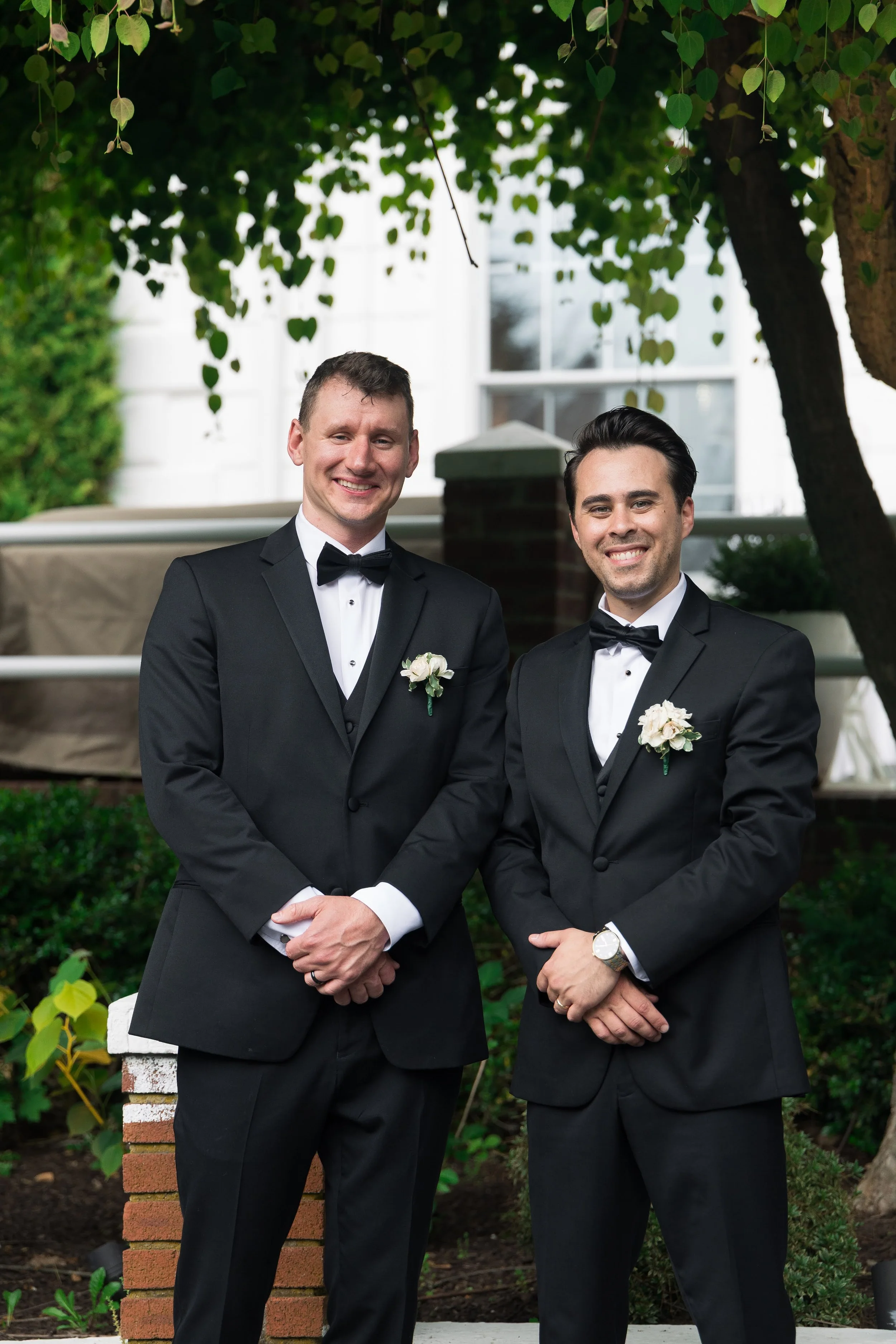 Two men in tuxedos with white shirts and black bow ties standing outdoors under a tree with green leaves, smiling at the camera.