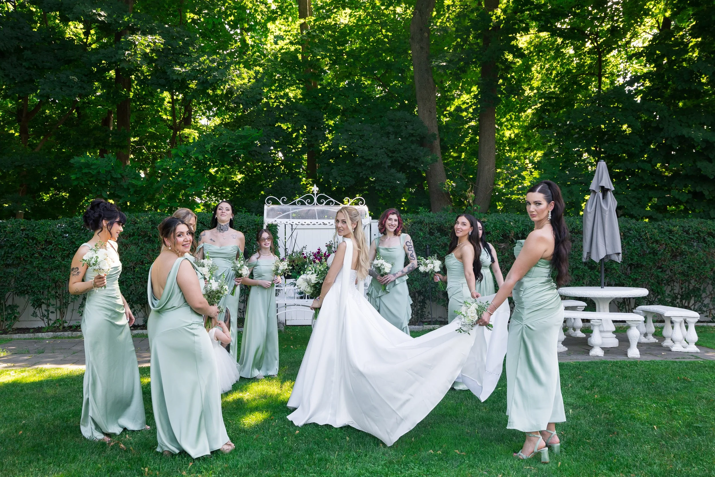 A group of women dressed in matching light green bridesmaid dresses holding bouquets and standing outdoors on grass during a wedding. The bride, in a white gown, is in the center. Tall trees with green leaves form the background.