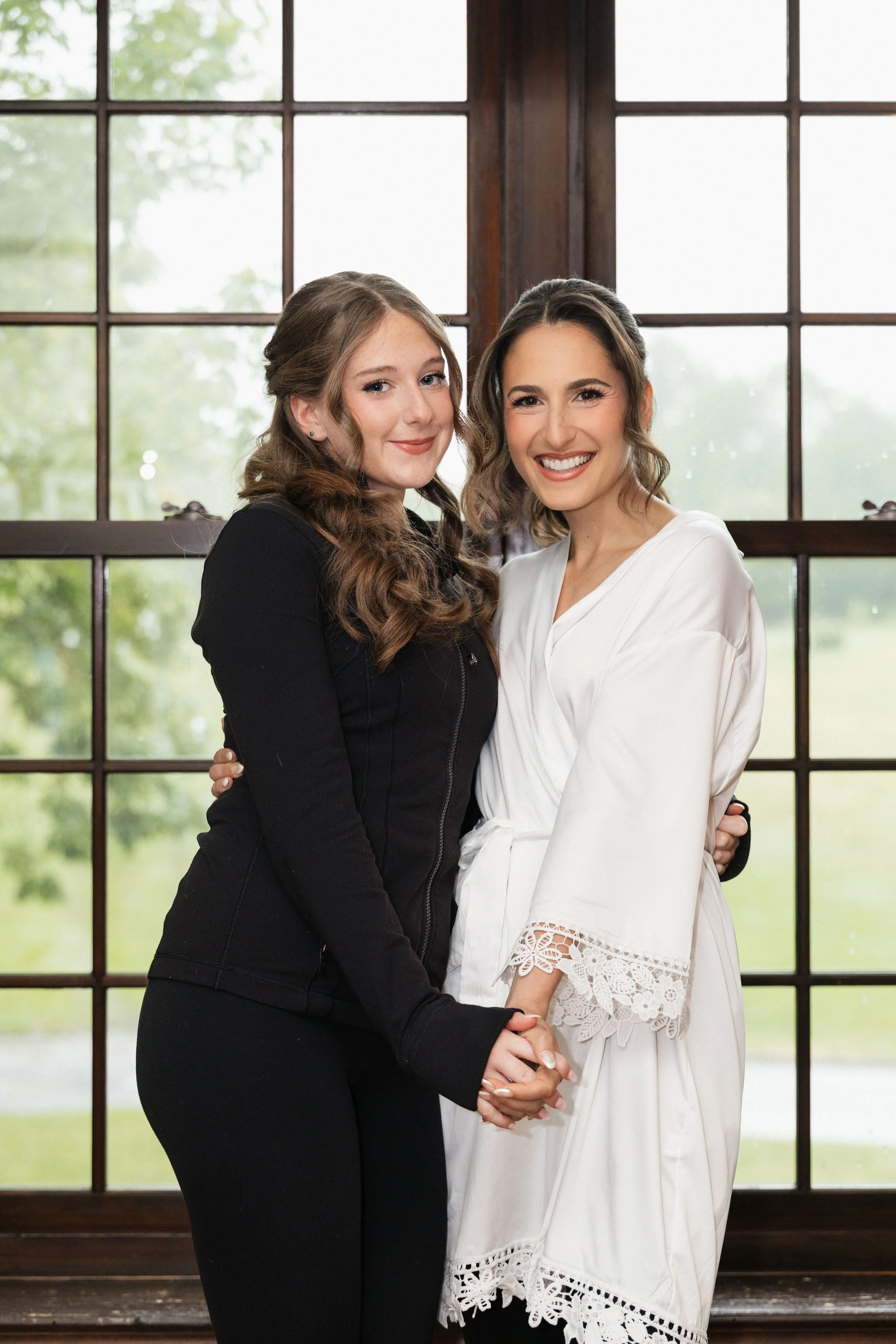 Two women standing inside in front of large window, holding hands and smiling.