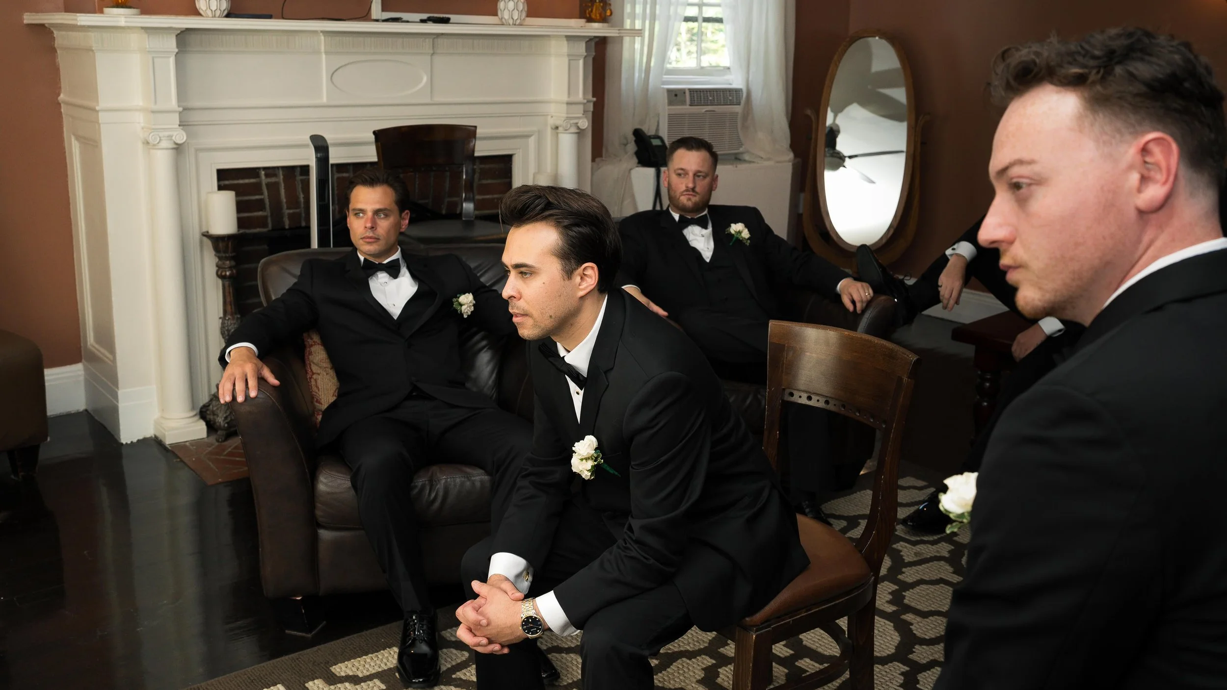 Four men in tuxedos with white boutonnières sitting in a living room, some looking serious or pensive, with a fireplace, mirror, and window behind them.