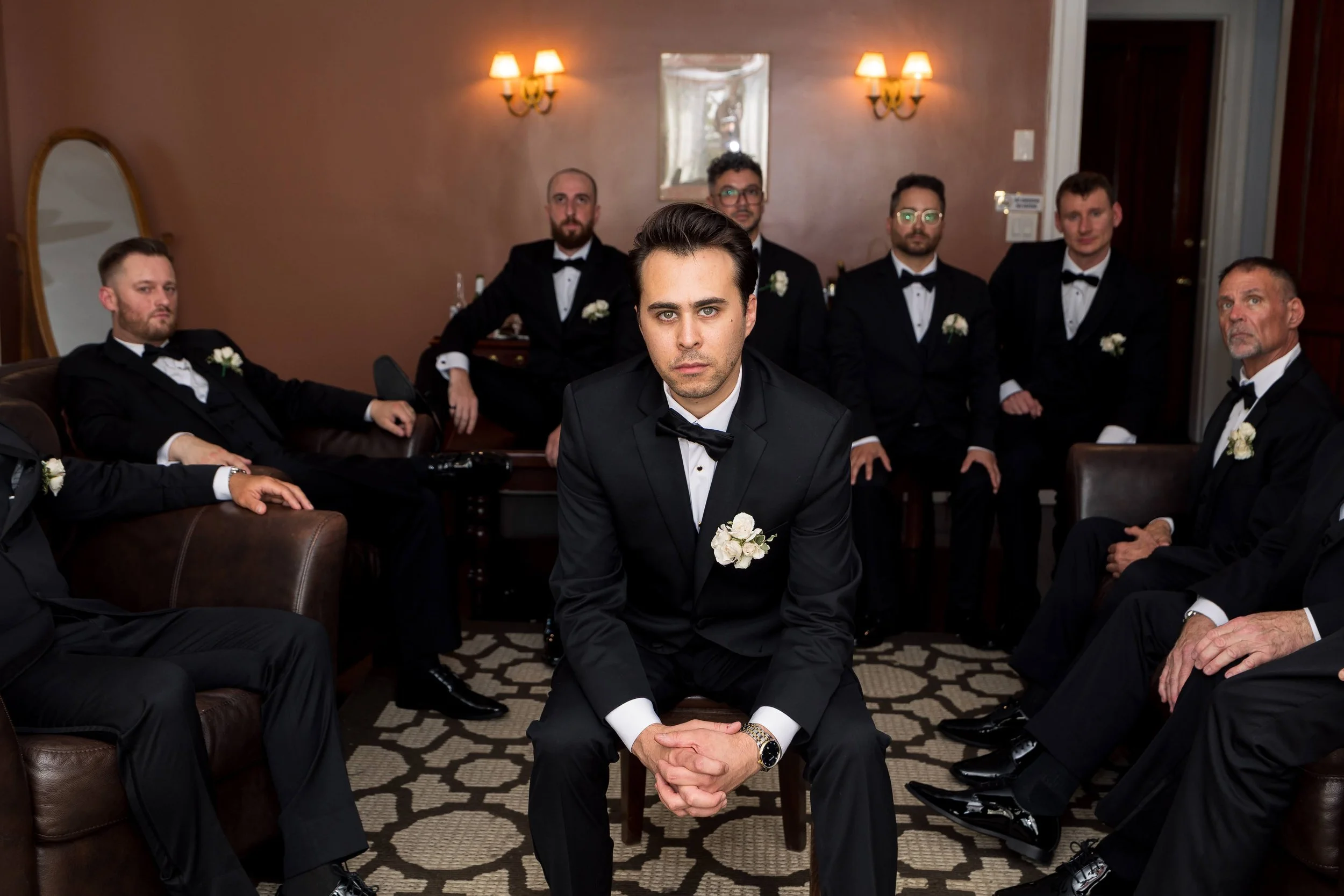 A group of seven men dressed in black tuxedos with white dress shirts and black bow ties, sitting and kneeling in a formal room. The man in the center, kneeling, has dark hair and a boutonniere, holding his hands together with a serious expression. T