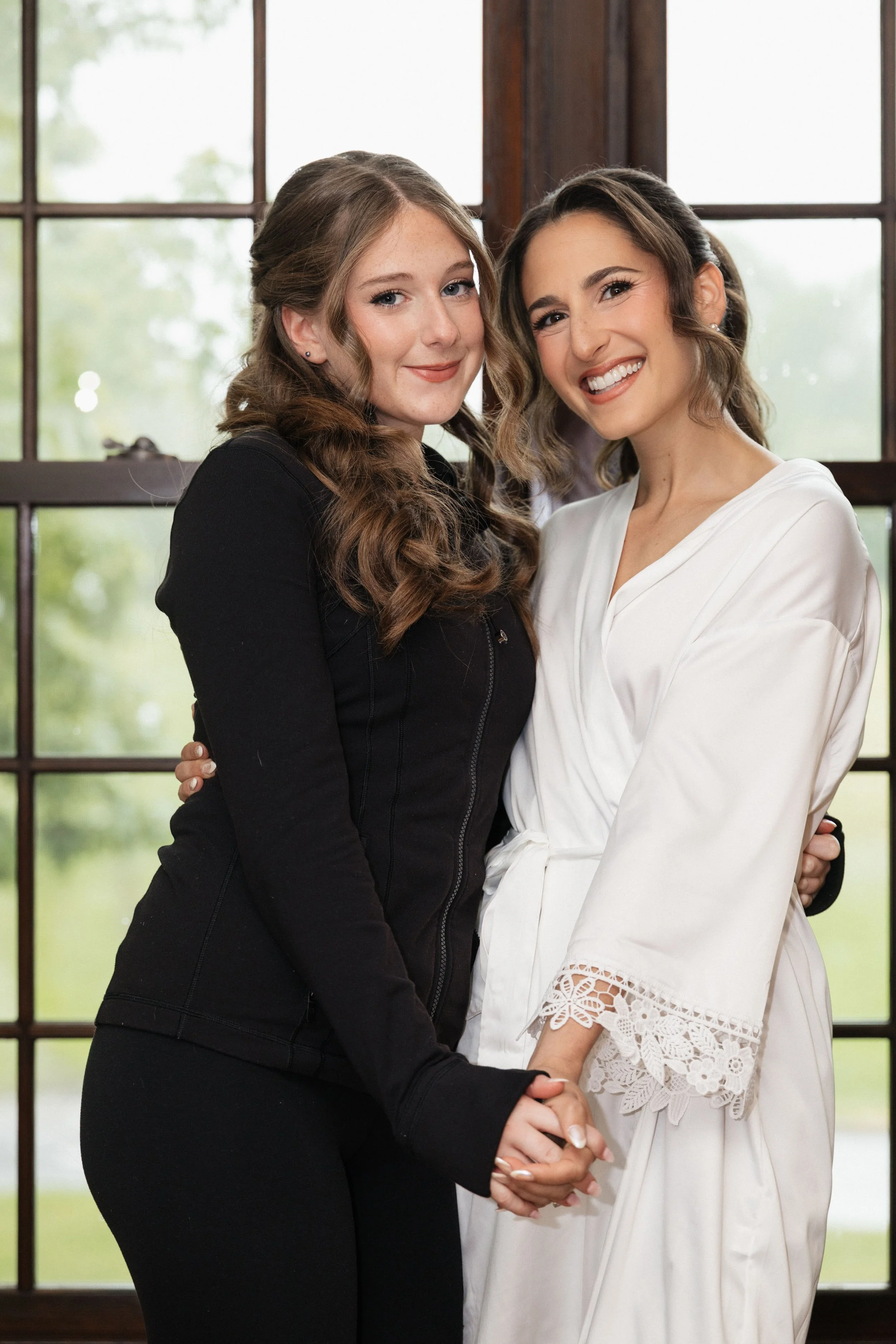 Two women, one with long brown hair in a black outfit and the other with shoulder-length dark hair in a white outfit, holding hands and smiling in front of windows with green trees outside.