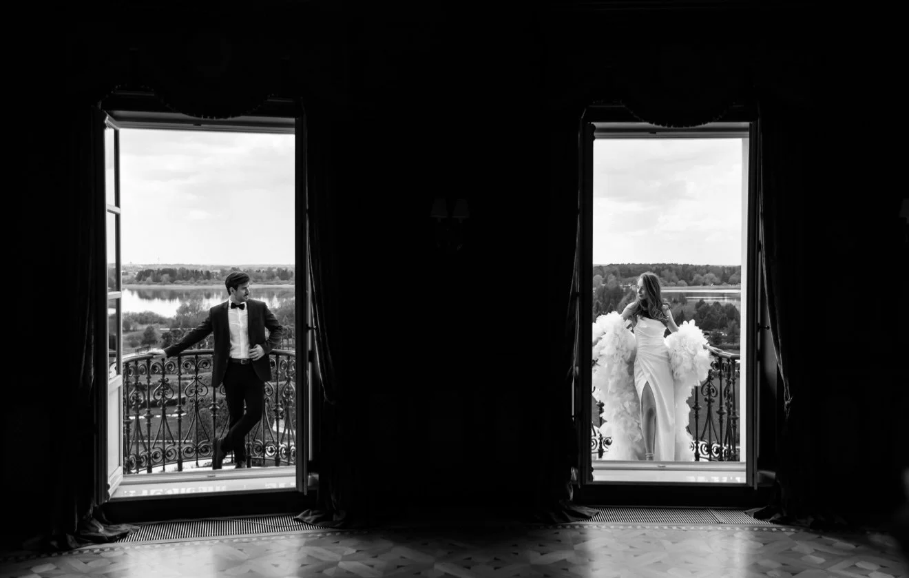 Black and white photo of a man in a tuxedo and a woman in a wedding gown standing on separate balconies, opening doors to view a scenic landscape with water and trees in the background.