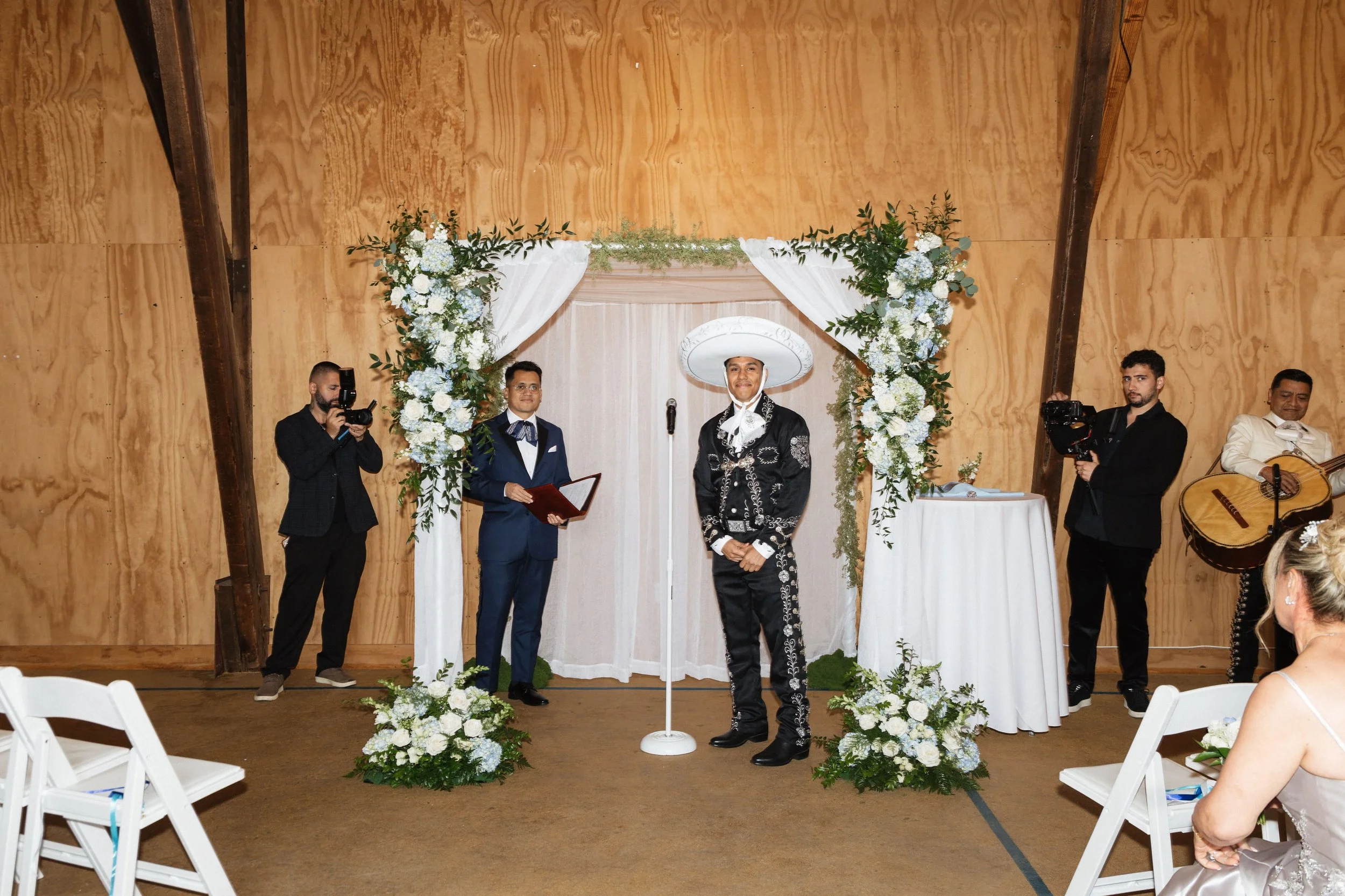 A wedding ceremony with an officiant in a black and white embroidered suit, a groom in a dark blue suit with a bow tie, and a person in a sombrero hat standing at a decorated white arch with floral arrangements, with musicians on either side, inside 