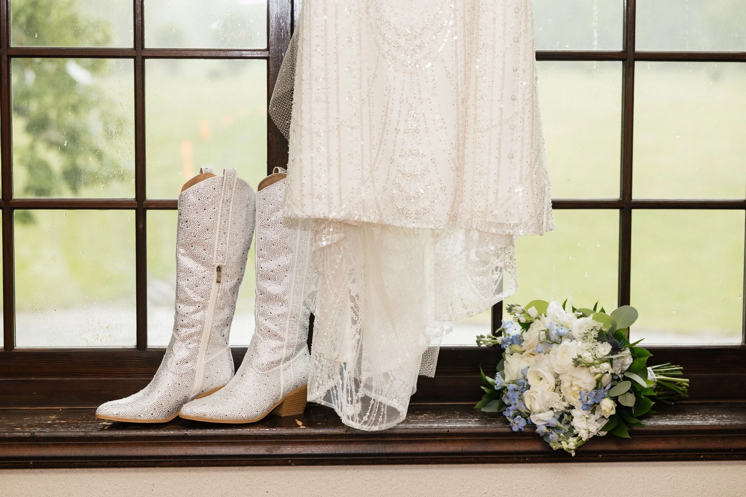 A window sill with a pair of white, glittery, knee-high lace boots, a white lace wedding dress, a bouquet of white and blue flowers, and a vase of white flowers inside on the window sill. The window has multiple panes and shows a blurred outdoor scen
