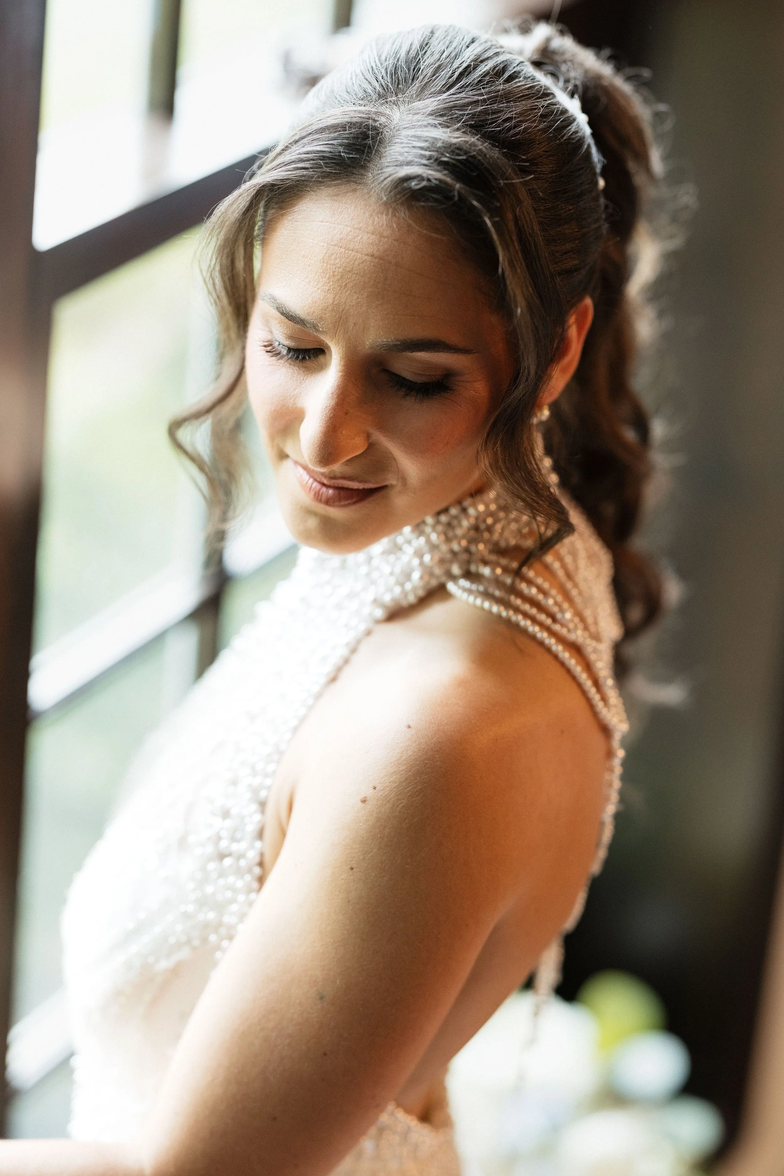 A woman with a floral headband and pearl dress posing with closed eyes near a window with sunlight.
