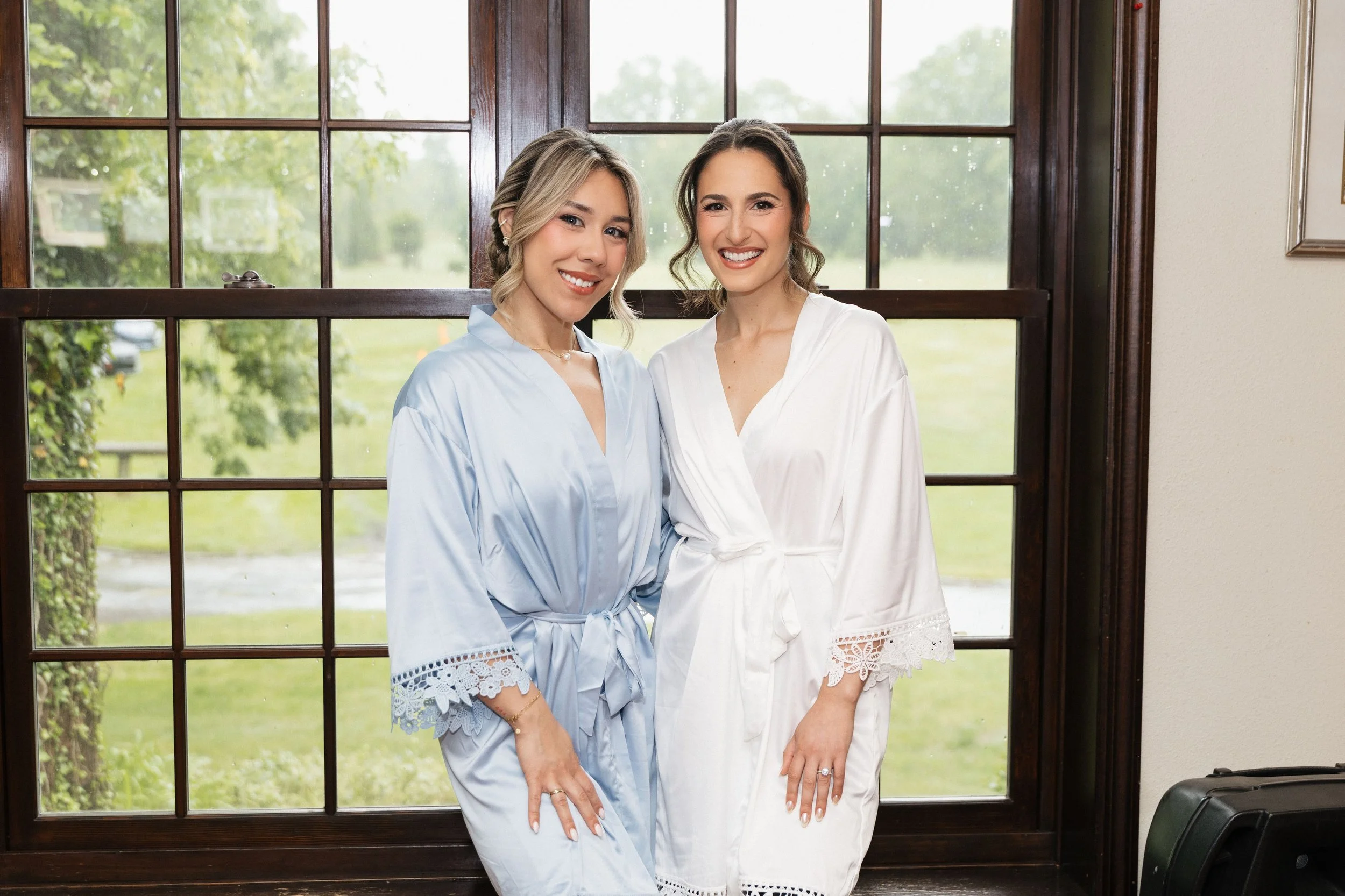 Two women standing side by side smiling in front of a large window with green trees outside. The woman on the left has blonde hair tied back, wearing a light blue robe with lace trim. The woman on the right has brown hair styled in loose waves, weari