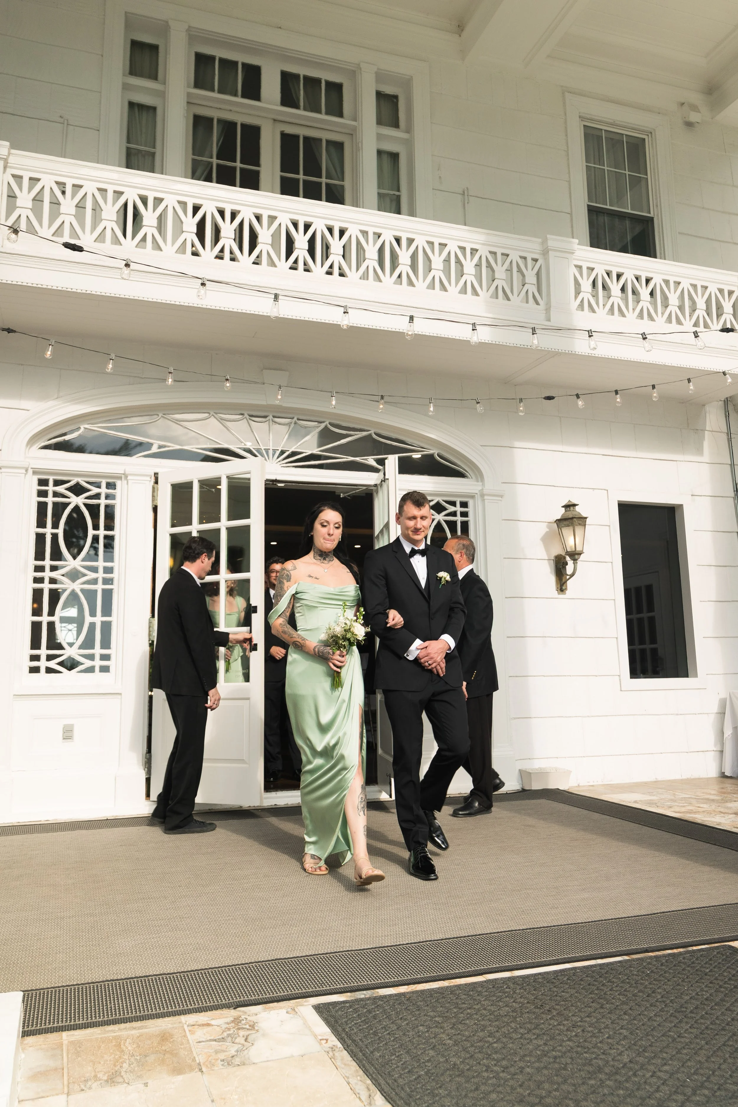 People dressed in formal attire leaving a white building with large windows and a balcony, during a special event or celebration.