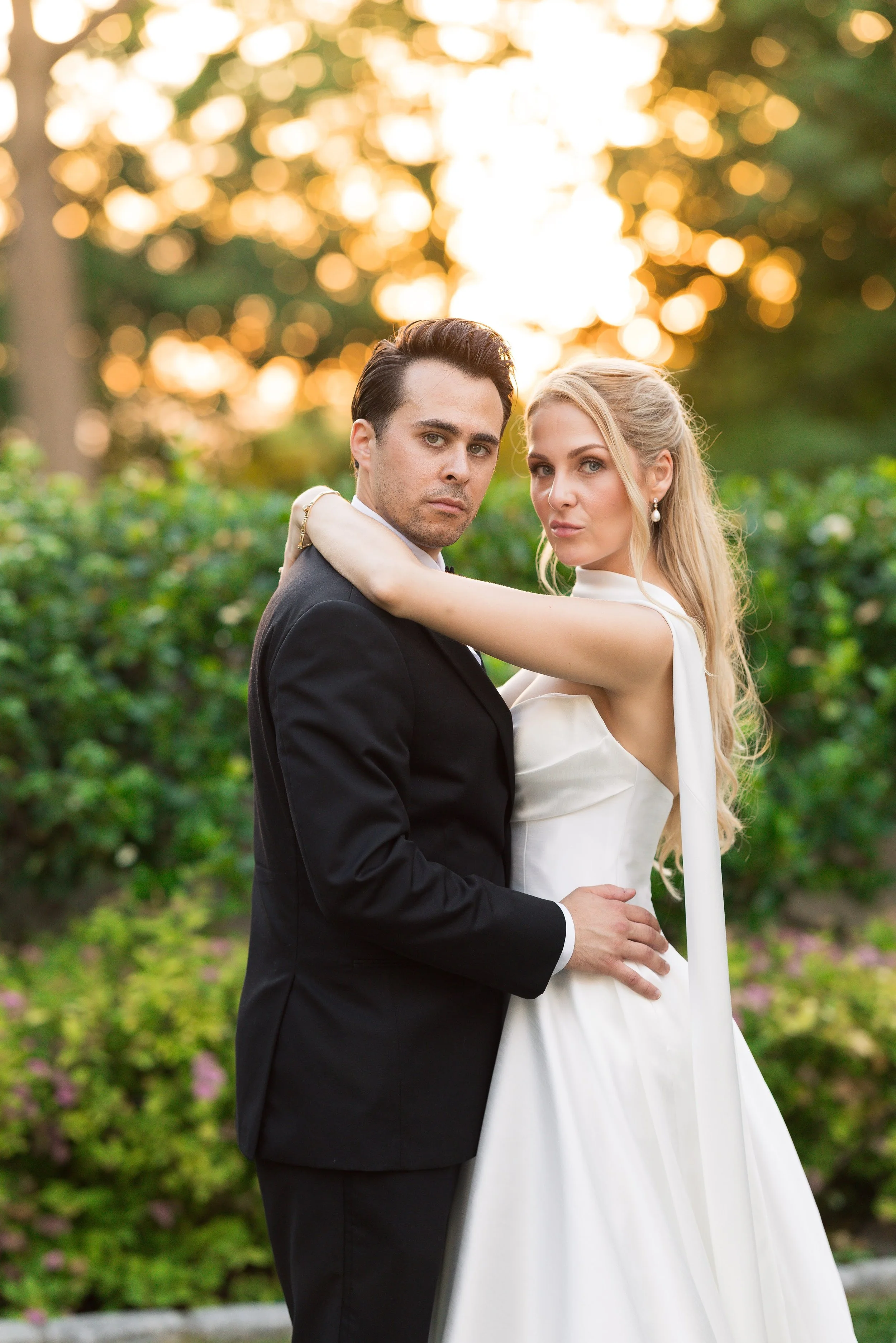 A man and woman in wedding attire standing outdoors at sunset, with trees and foliage in the background.