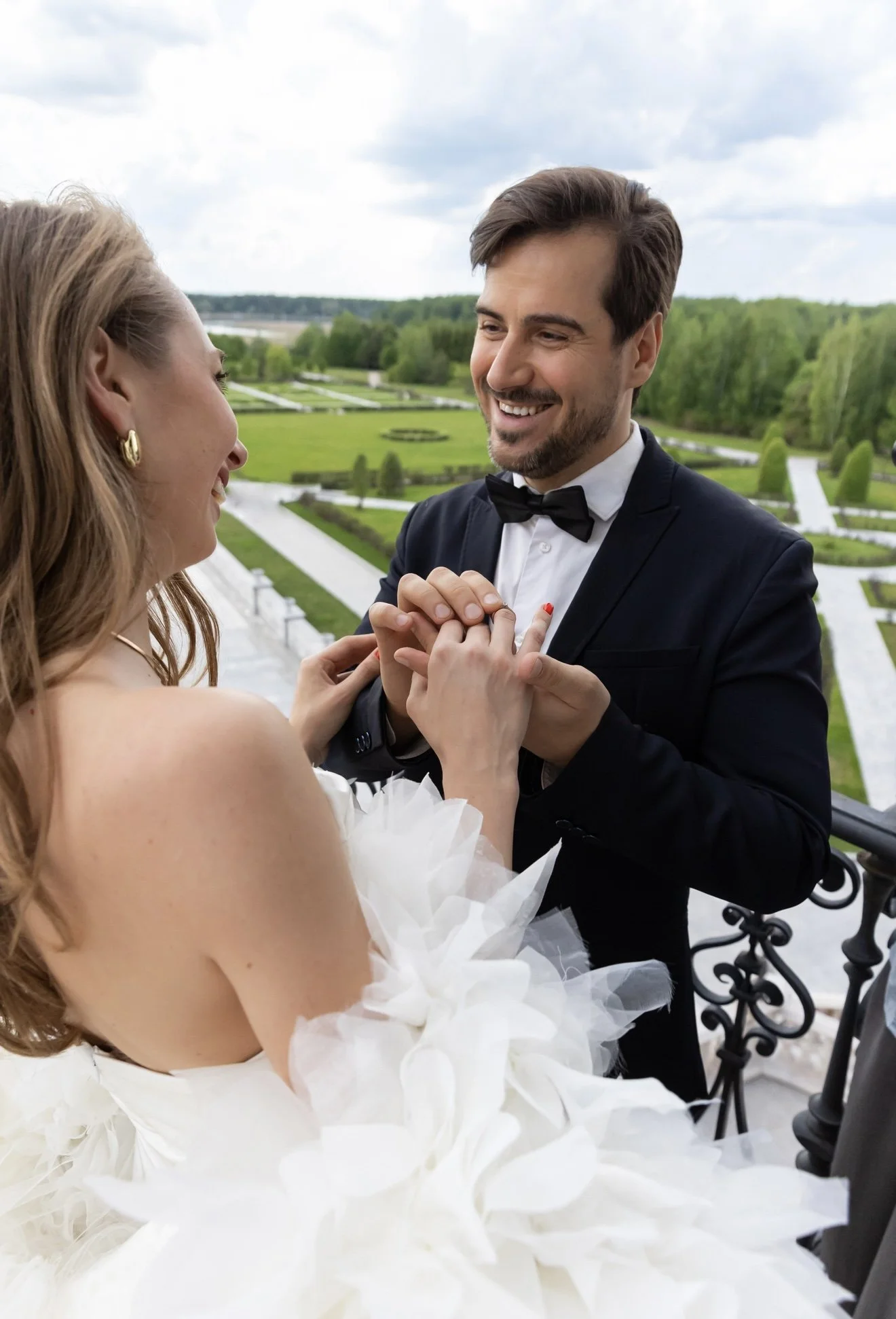 A bride and groom share a moment outdoors, holding hands and smiling at each other, with a lush green landscape and cloudy sky in the background.