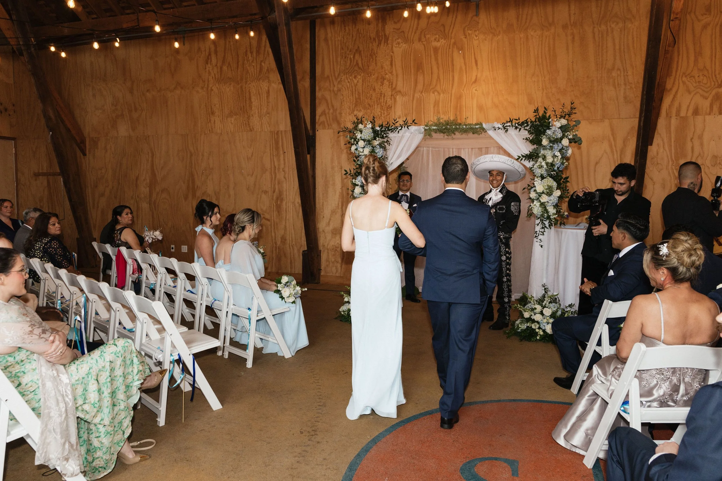 A wedding ceremony taking place in a rustic indoor setting with wooden walls. The bride and groom stand before an officiant under a floral arch. Guests are seated on white chairs, some holding bouquets. A photographer and videographer are capturing t