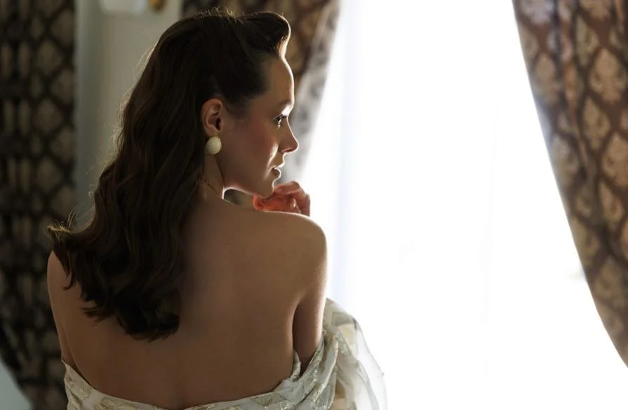 Woman with wavy brown hair and pearl earrings, facing a window with ornate curtains, wrapped in a white shawl or dress.