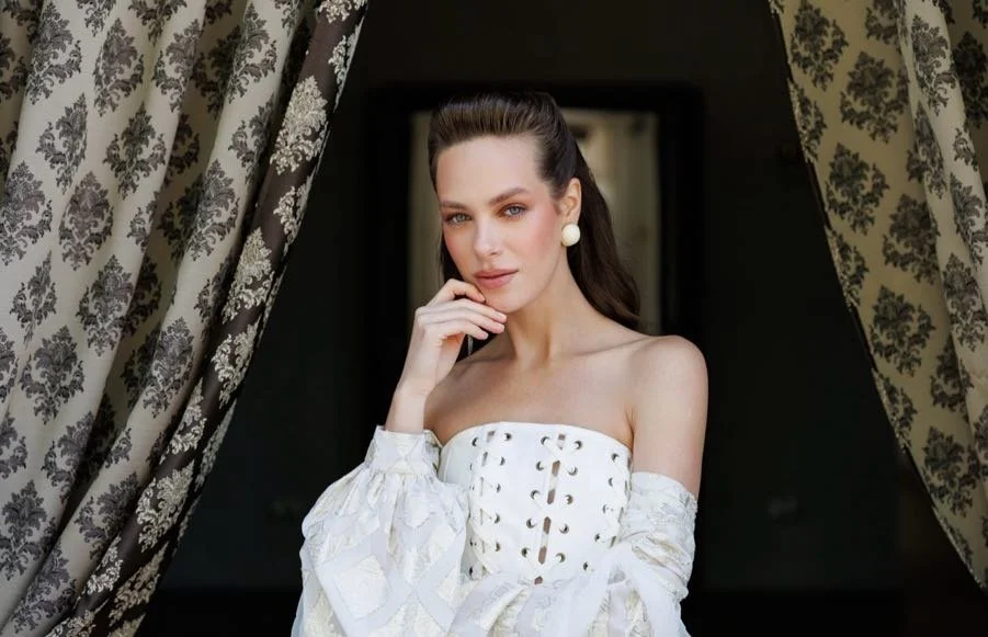 A woman with long brown hair wearing a white strapless dress with cut-out details and a matching off-the-shoulder sleeve, standing between patterned curtains in a dark room.