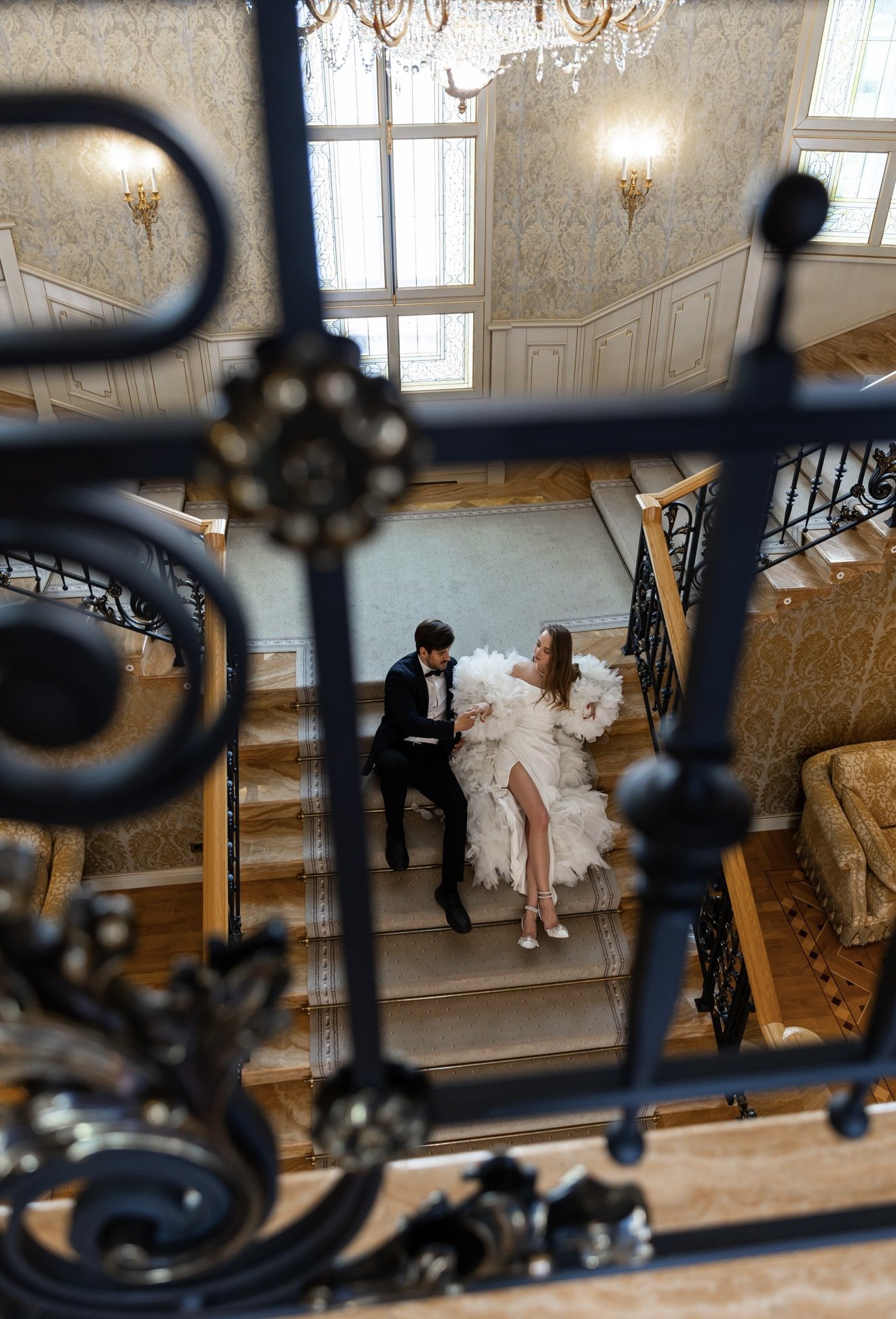 A man and woman dressed in formal wedding attire sitting on a staircase, seen through an ornate black metal railing with gold accents, in a luxurious interior with wallpapered walls, chandeliers, and large windows.