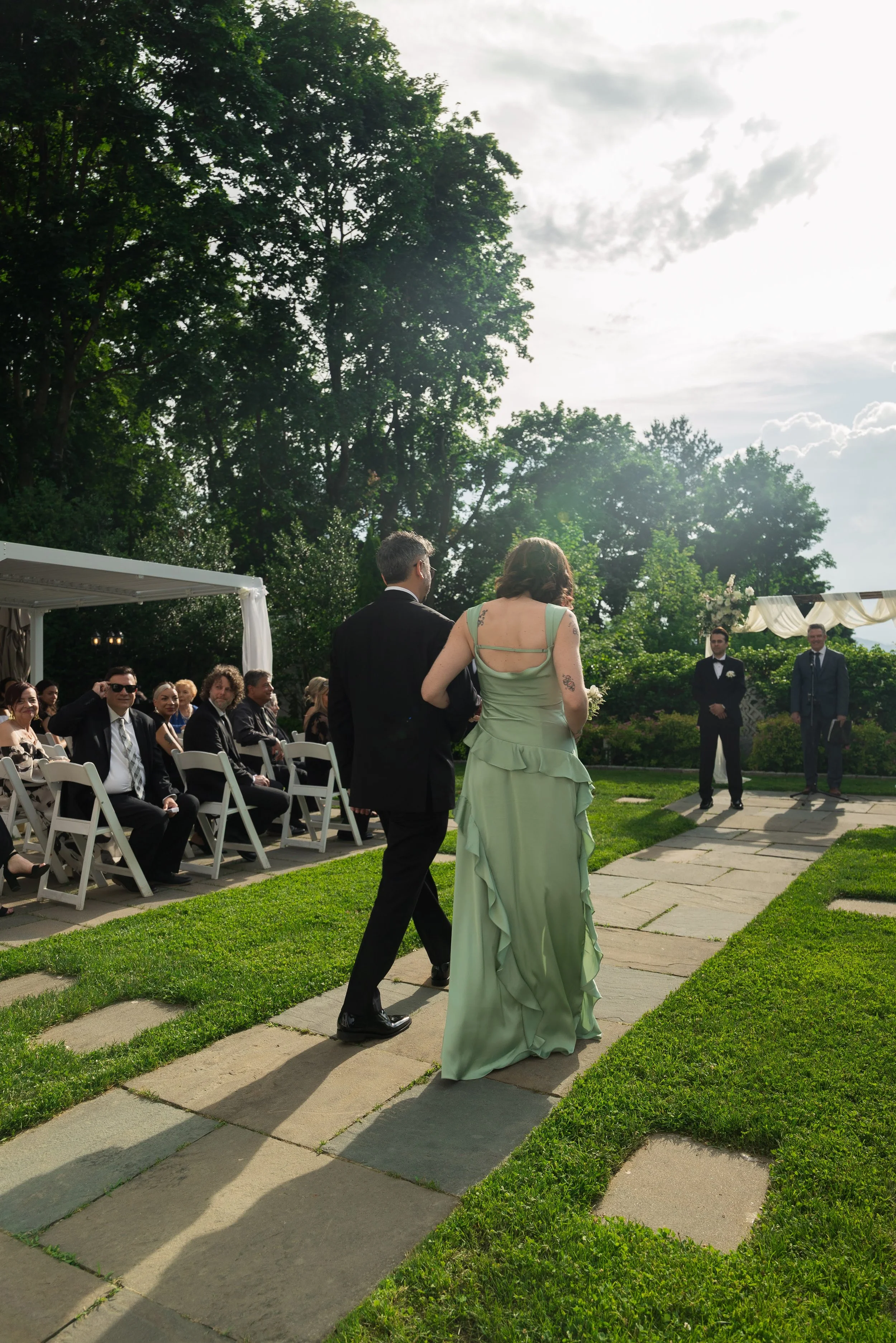 A wedding ceremony outdoors on a sunny day with a bride in a pastel green dress and a groom in a black tuxedo walking on a stone pathway. Guests are seated on white chairs under a white canopy, and a background of lush green trees and a cloudy sky