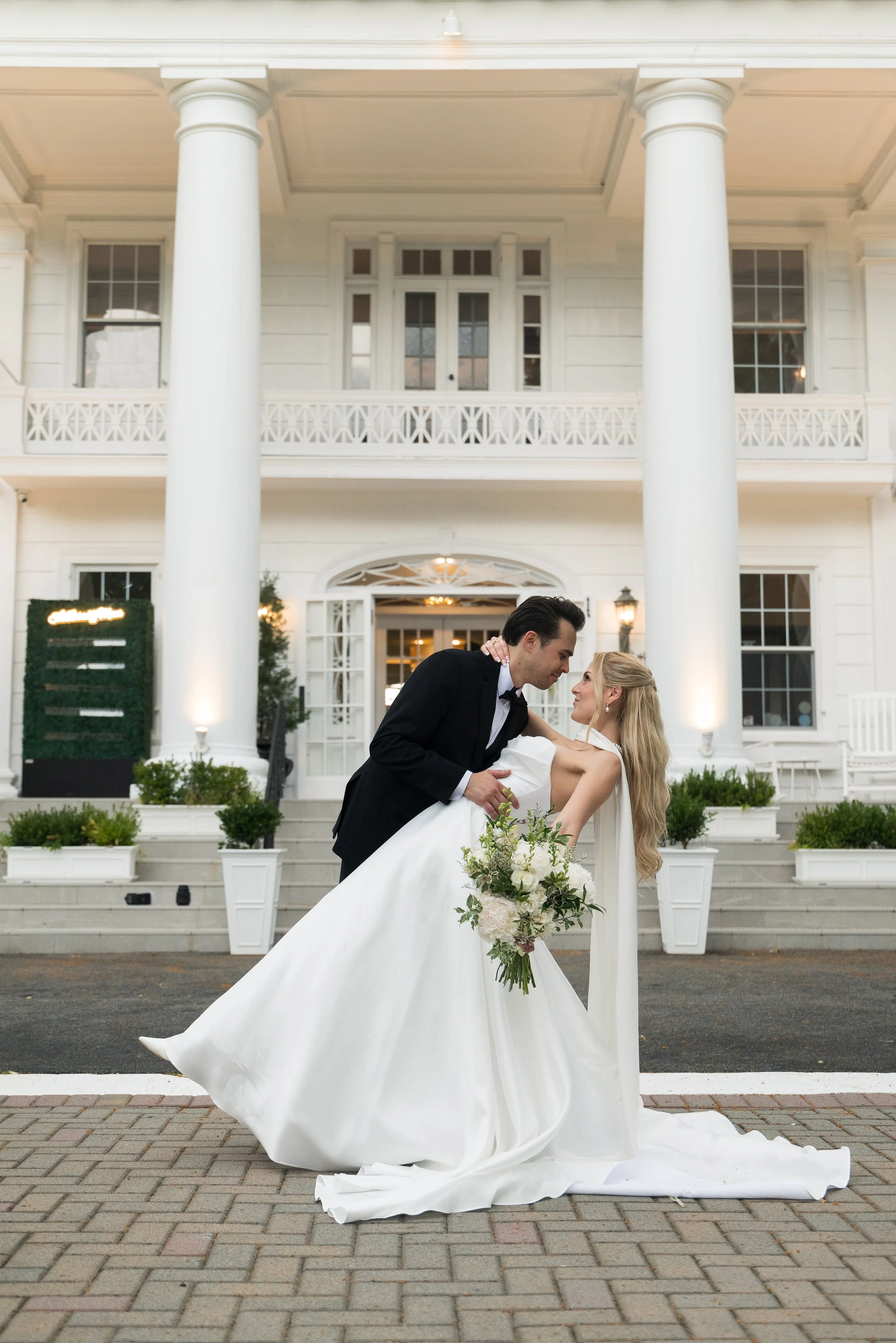 A bride and groom sharing a dance outside a white mansion during their wedding. The groom is dressed in a black tuxedo, and the bride is wearing a white wedding gown and holding a bouquet of flowers. The groom is leaning over and smiling at the bride