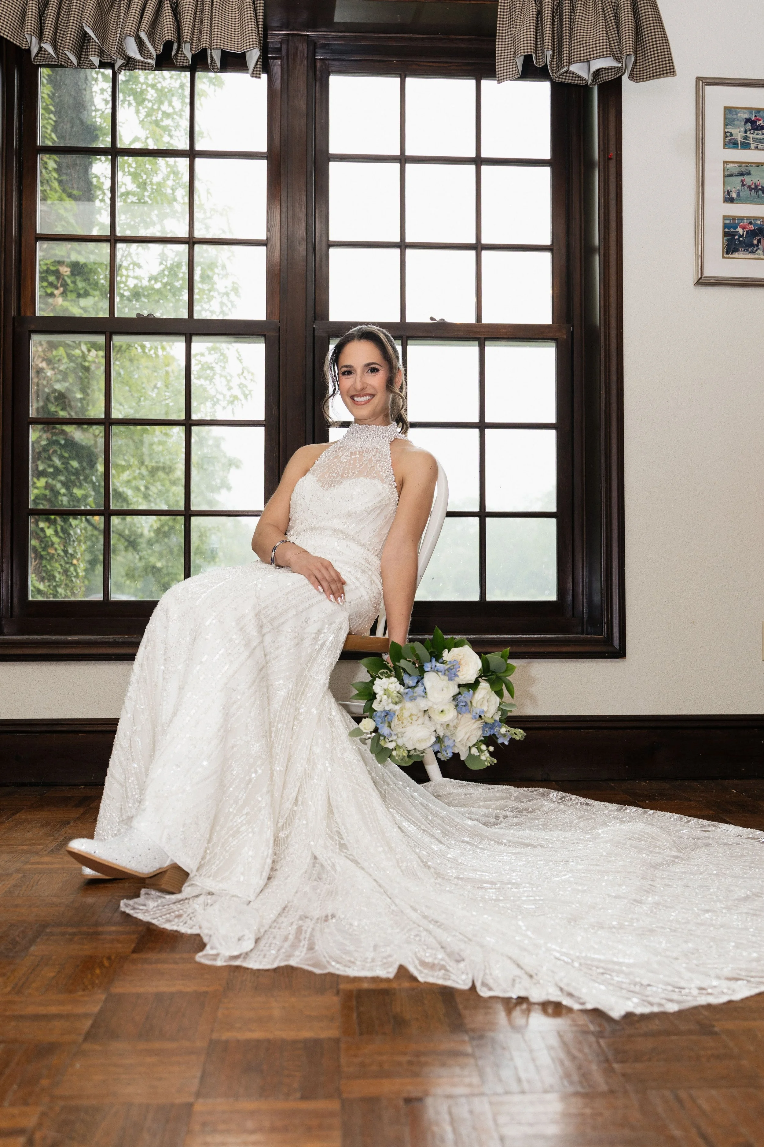 A smiling woman wearing a white wedding dress sitting on a white chair by a large window with dark wood trim and a plaid valance, holding a bouquet of white and blue flowers on the floor.