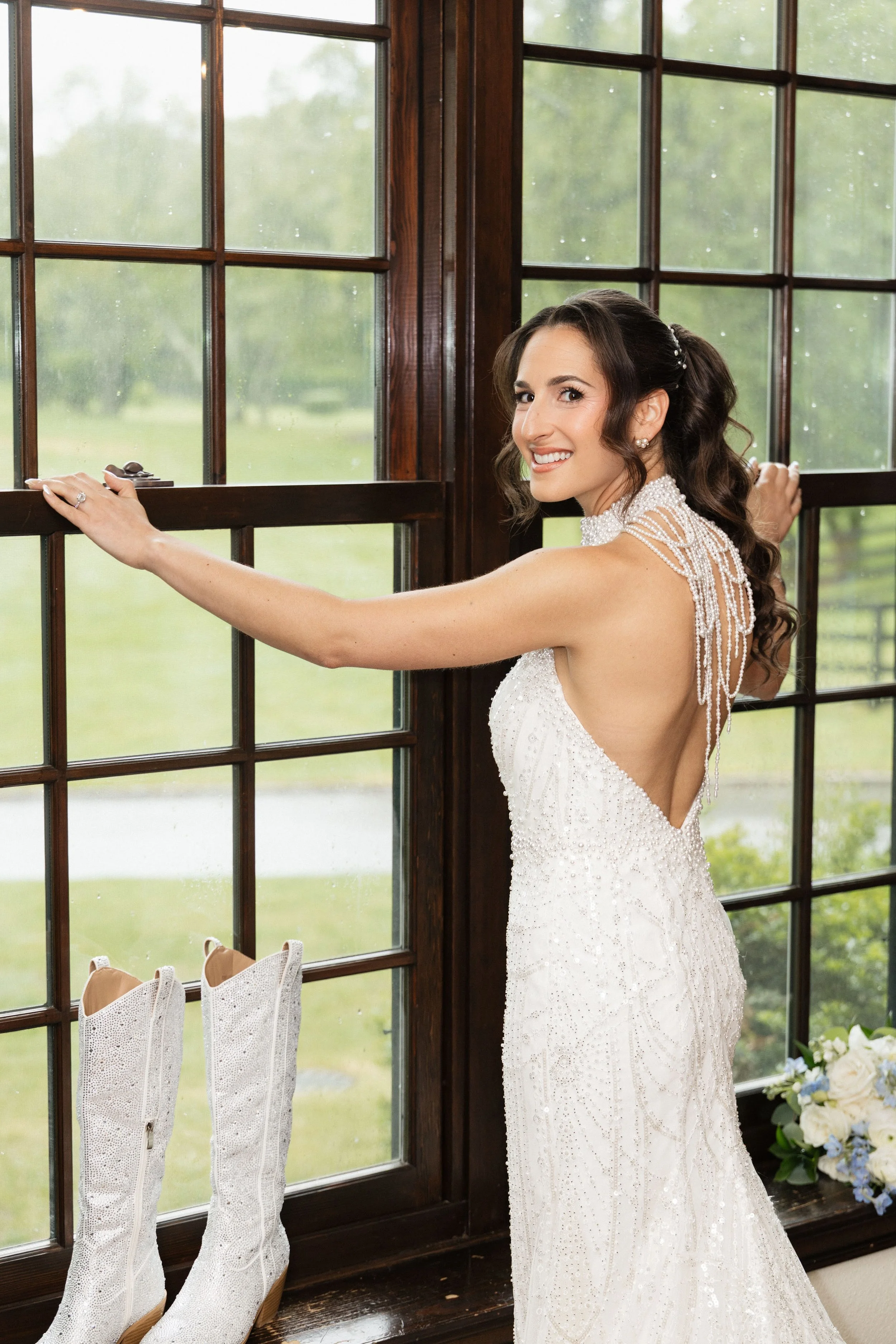 A bride in a white wedding gown with pearl details, standing by a large window with wooden frames, smiling and looking over her shoulder. There are white cowboy boots on the windowsill and a bouquet of white roses with greenery nearby.