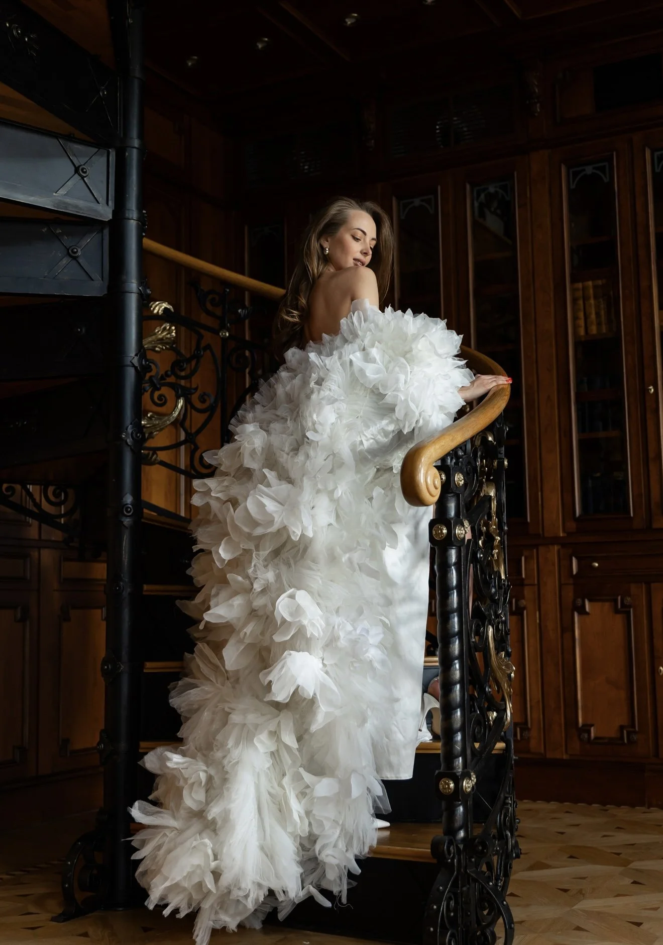 A woman in a white, ruffled, strapless gown standing on a staircase in a luxurious room with dark wood paneling and bookshelves.