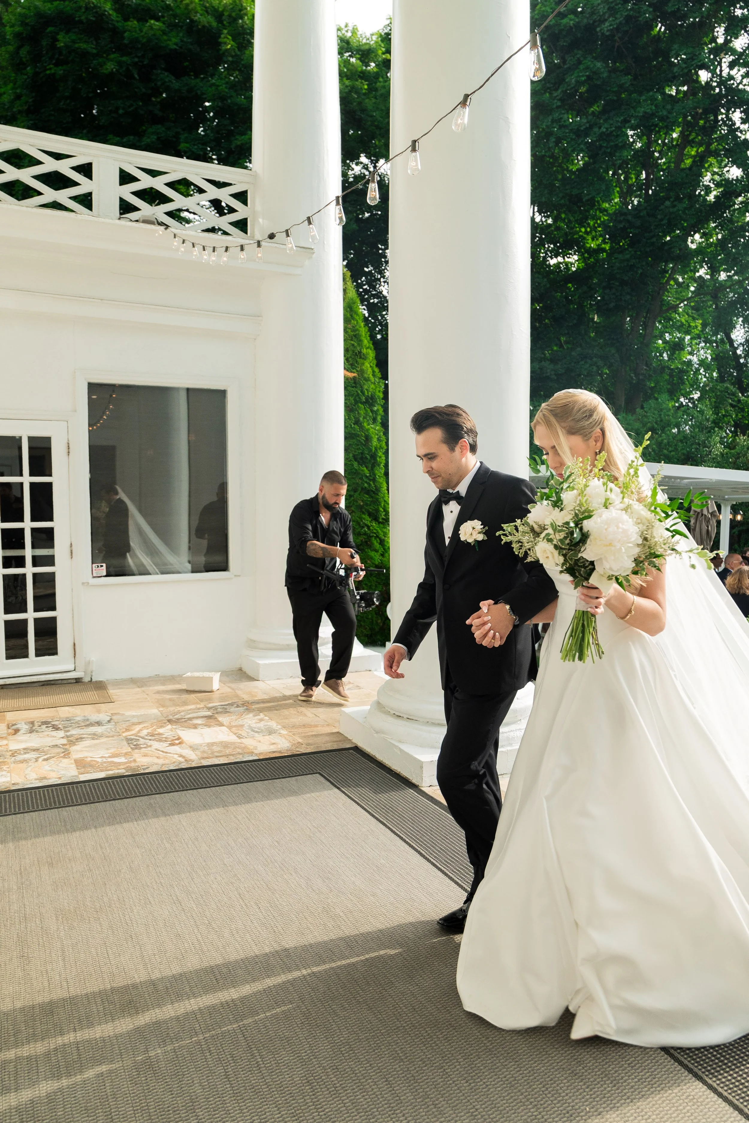 A bride holding a bouquet of white flowers and greenery walking with a groom in a black tuxedo outside a white building with large pillars. A person is standing nearby, adjusting something on a bicycle. The setting includes string lights and green tr