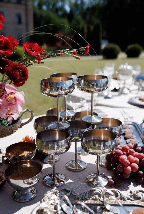 A table set outdoors with silver dessert cups, some filled with spices, alongside a bunch of grapes and a floral arrangement of red and pink flowers.