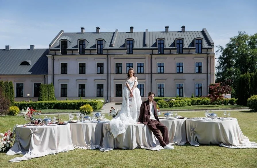 Bride in a wedding dress standing on a table beside a groom in a suit, set outdoors in front of a large mansion with tables arranged for a celebration.