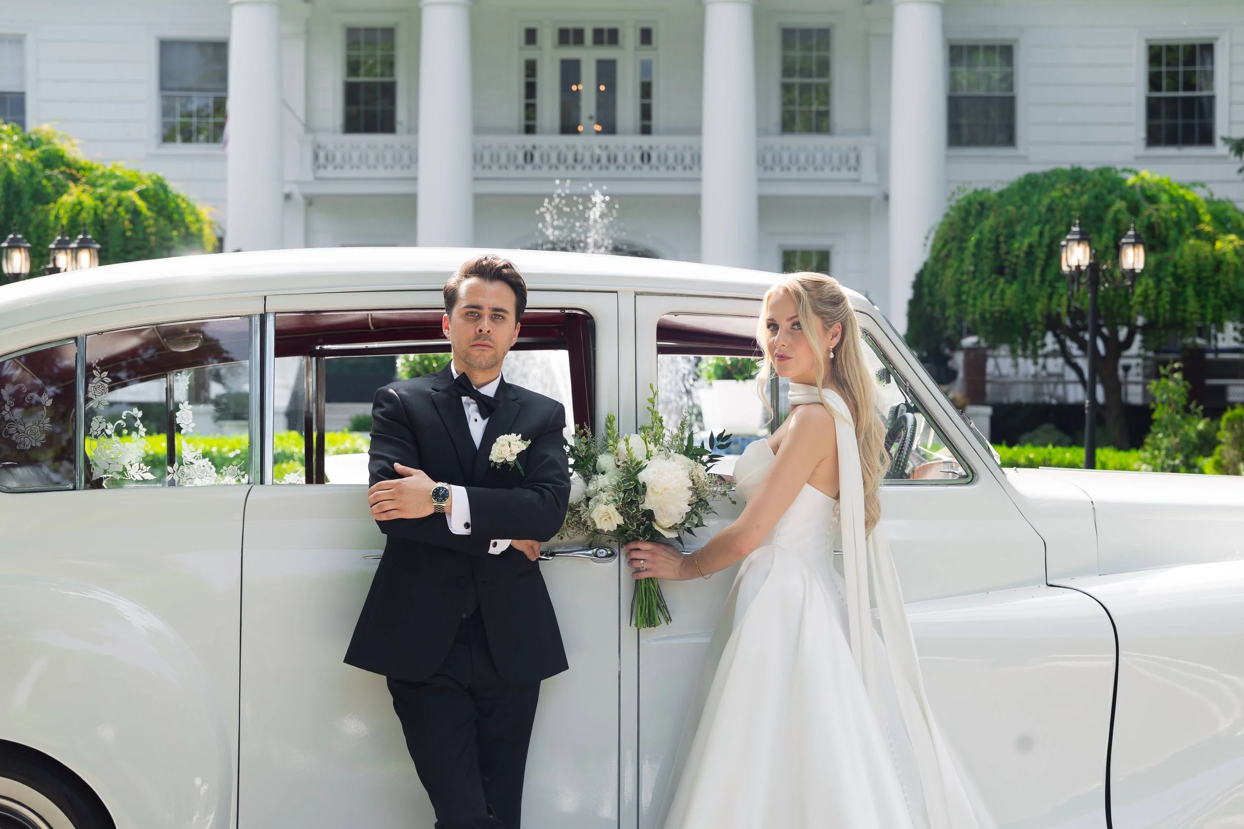 A bride and groom standing near a white vintage car in front of a large white house. The groom is wearing a black tuxedo with a bow tie and a white boutonniere, while the bride is in a white wedding gown holding a bouquet of white flowers. They are p