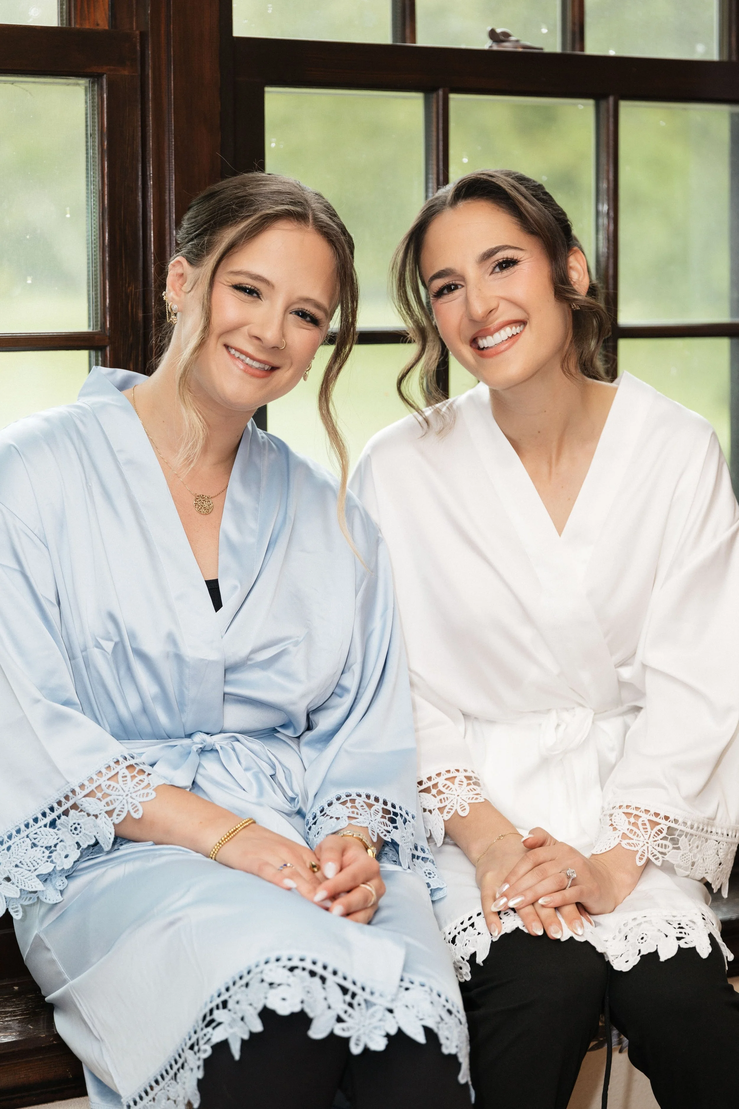 Two women sitting side by side, smiling, in front of a window with wooden framing. They are wearing lace-trimmed clothing, one in light blue and the other in white, with jewelry and styled hair.