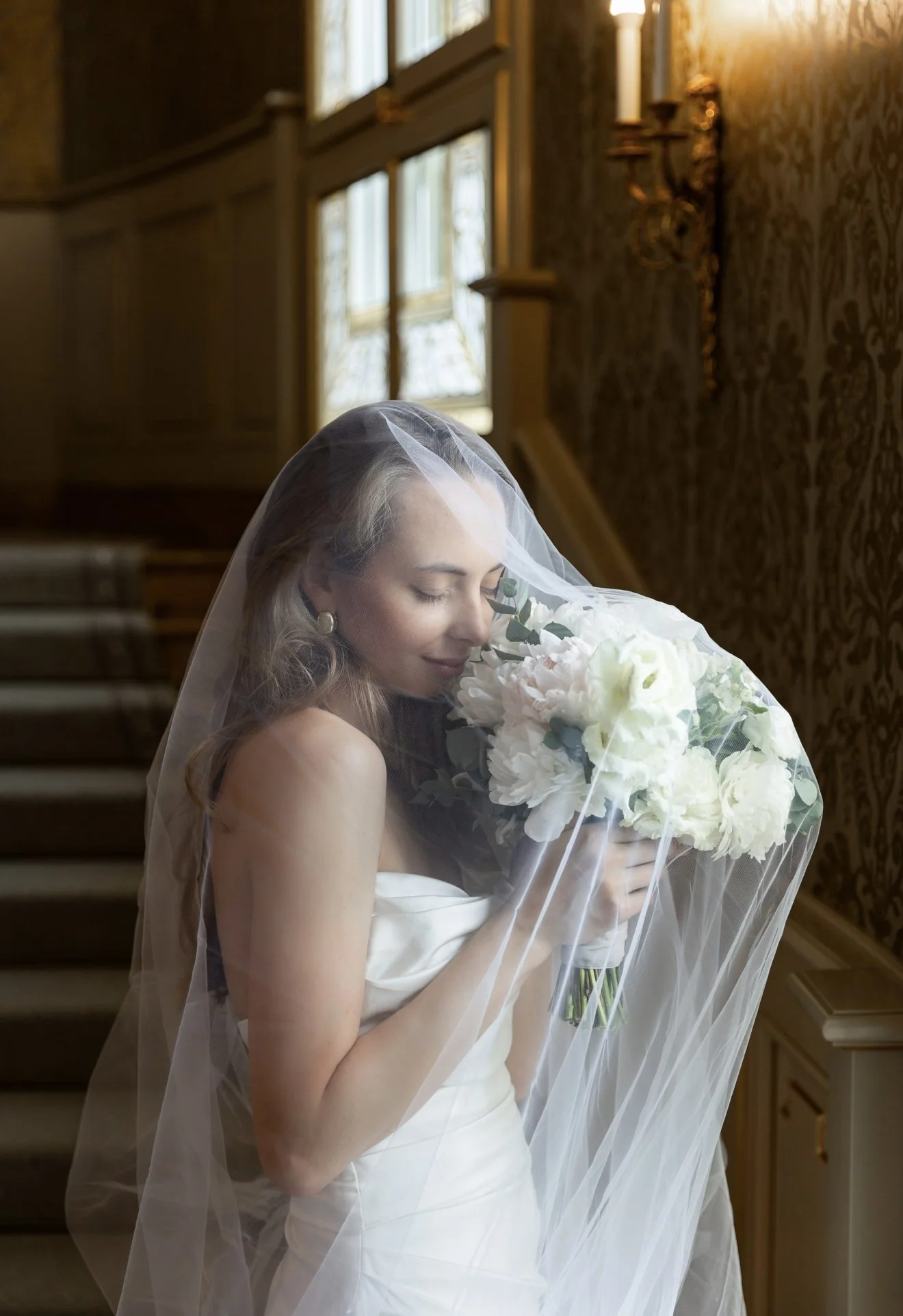 A woman in a wedding dress with a veil holding a bouquet of white flowers, standing in an elegant room with warm lighting and wooden details.