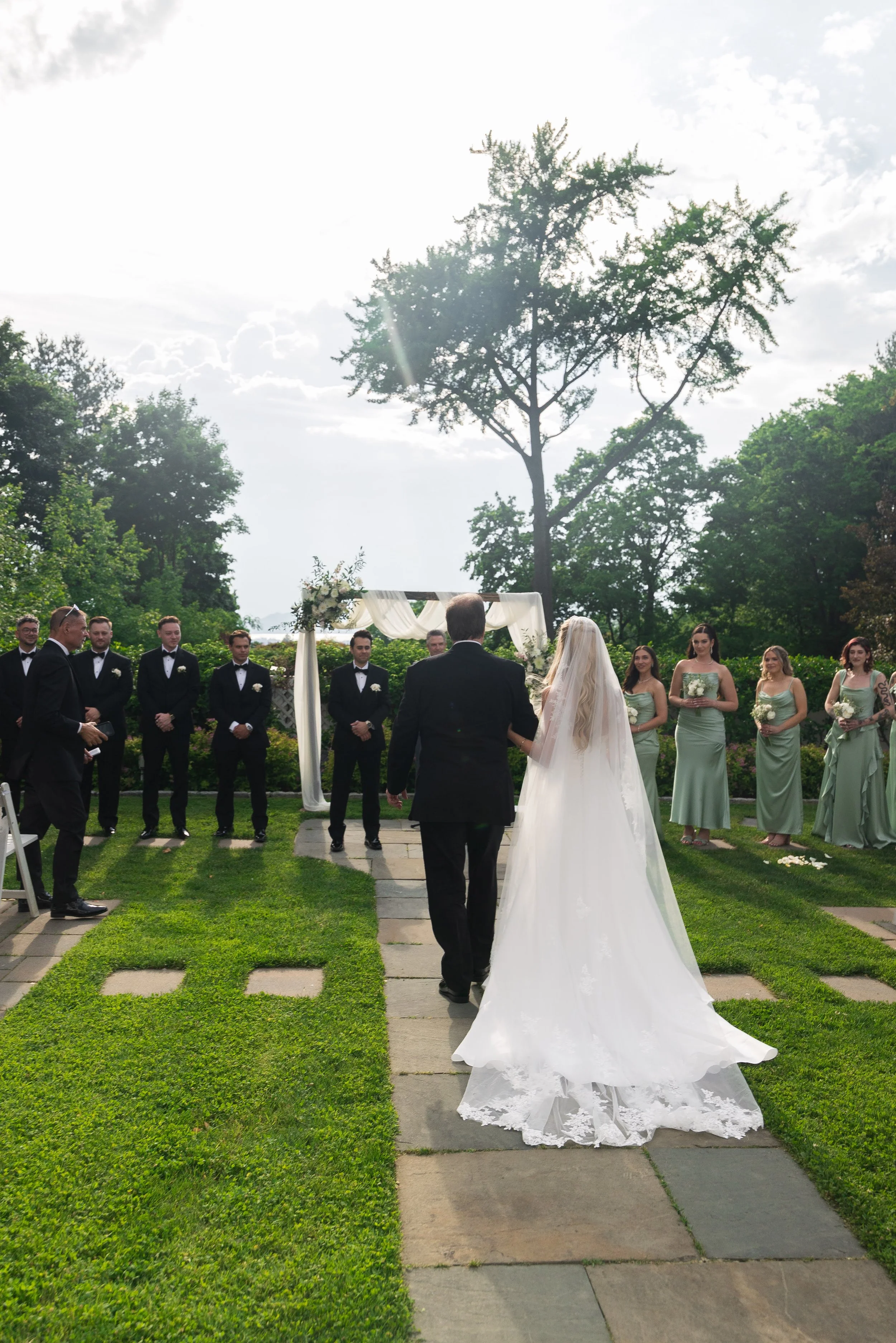 A wedding ceremony taking place outdoors with a bride and groom standing in front of a group of bridesmaids and groomsmen. The scene is set in a lush green garden with trees and a bright sky.