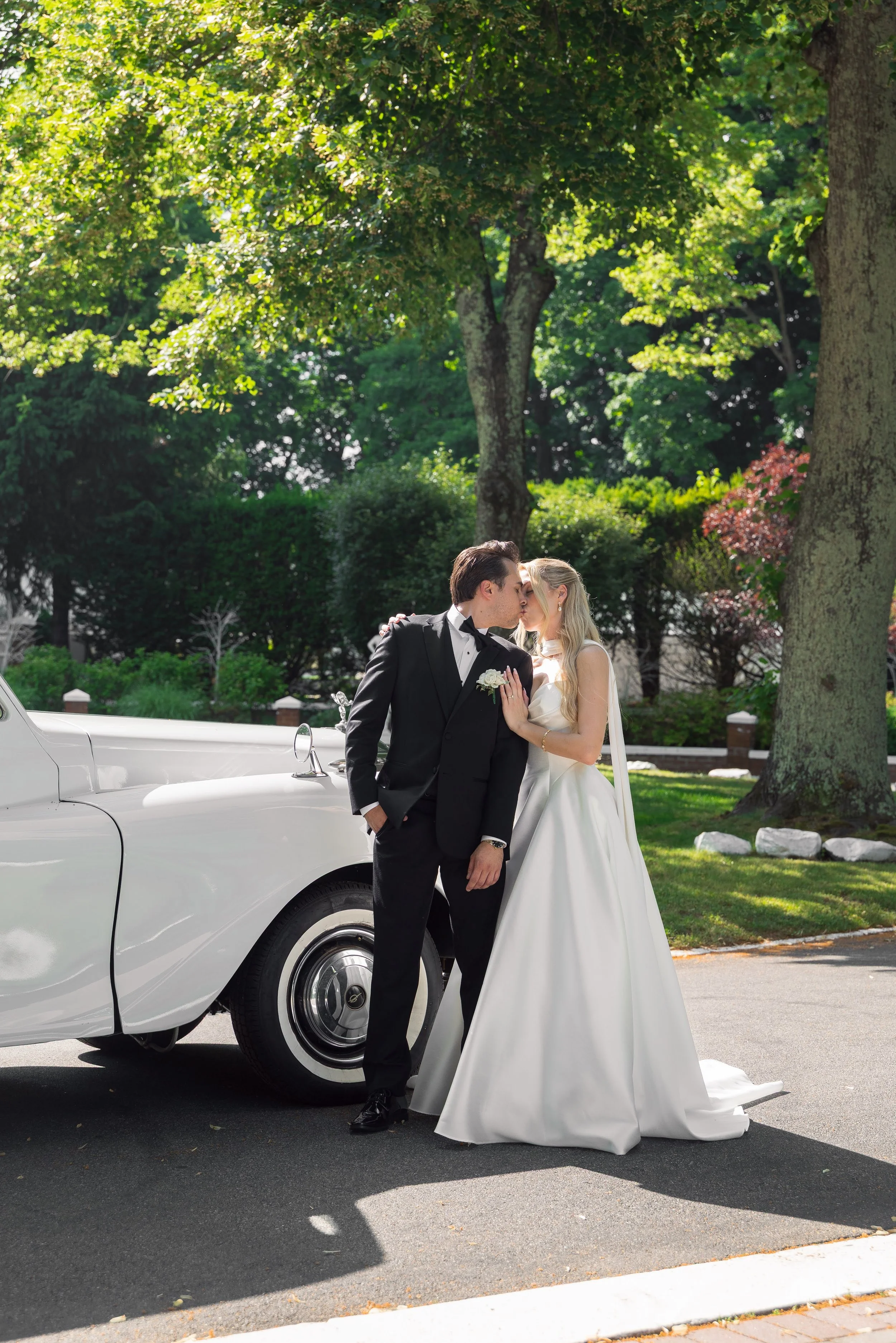 A bride and groom sharing a kiss next to a white vintage limousine in a park.