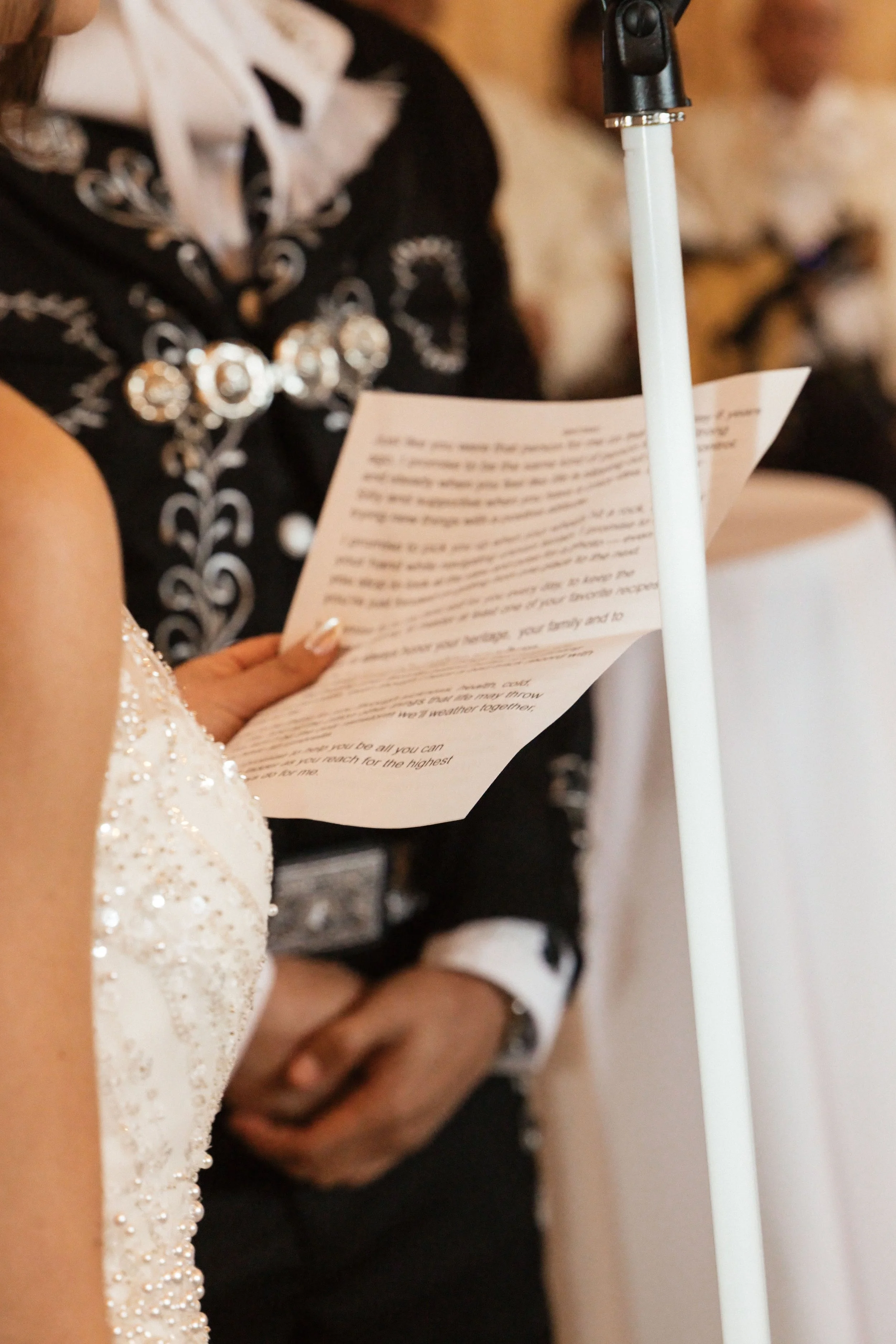 A woman reading a paper at a formal event, dressed in a white, beaded dress with pearl details, standing near a microphone stand.