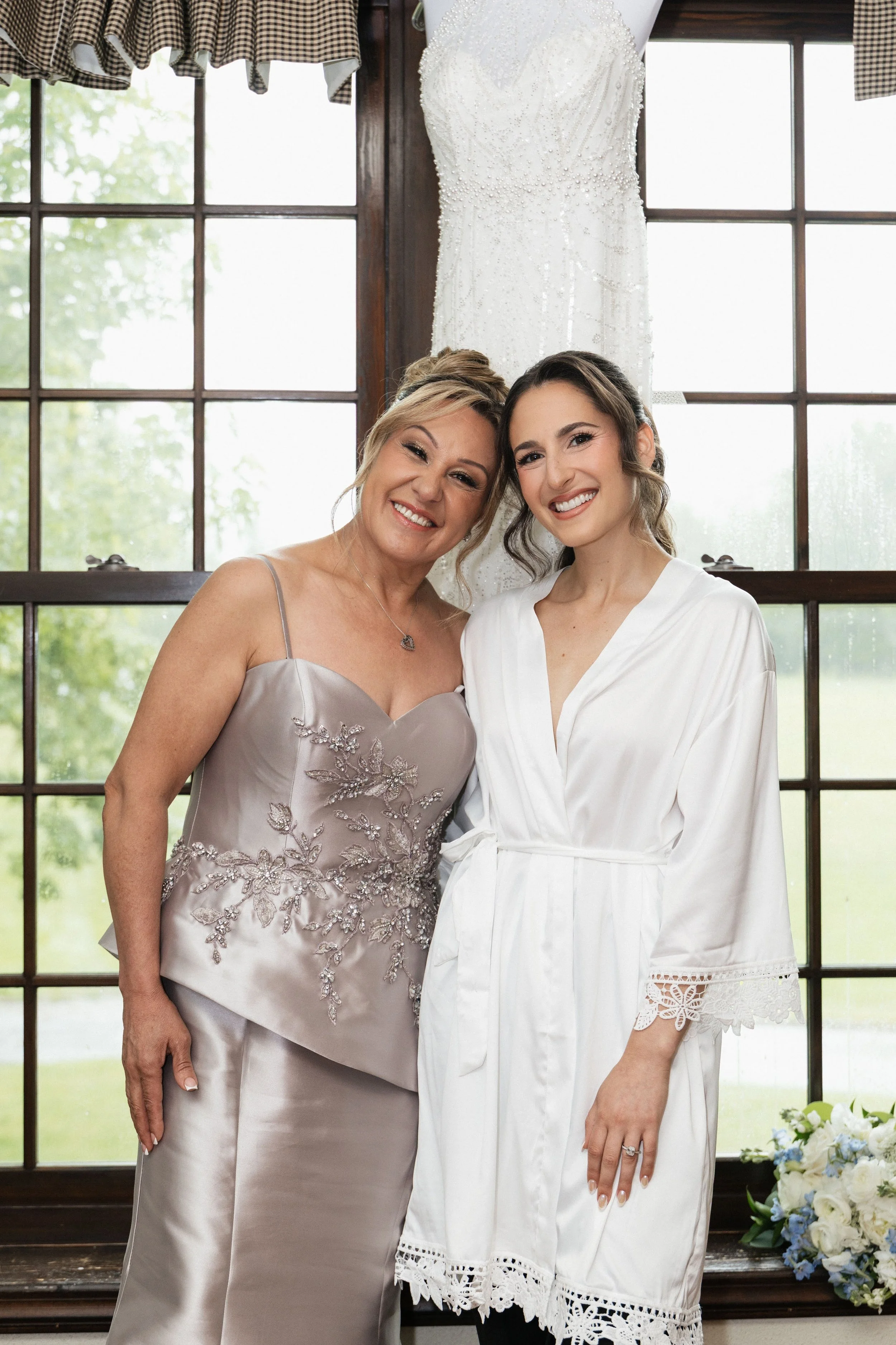 Two women, one older and one younger, smiling and standing close together in front of a large window with a white wedding dress hanging behind them. The older woman is wearing a satin dress with floral embroidery, and the younger woman is wearing a w