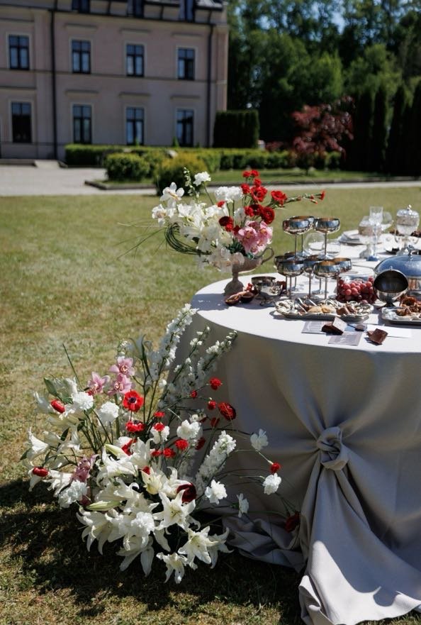 Elegant outdoor table setup with floral arrangements, glassware, and desserts on a white tablecloth, in a garden with a historic building and greenery in the background.