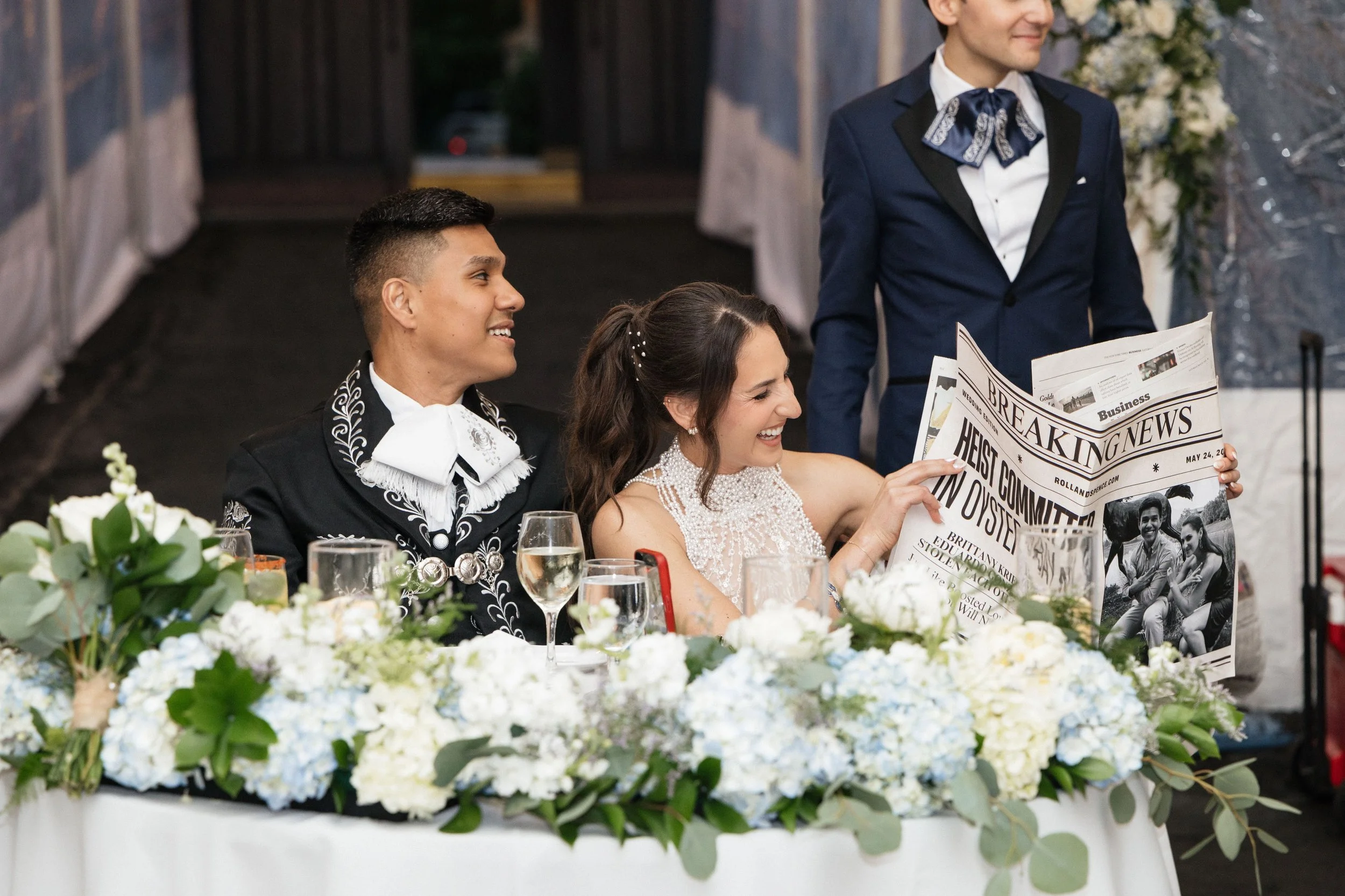 A wedding reception scene with the bride and groom seated at a table, smiling and looking at a newspaper. The table is decorated with white flowers and greenery, with glasses of champagne in front of them. A man in a blue suit is standing behind them