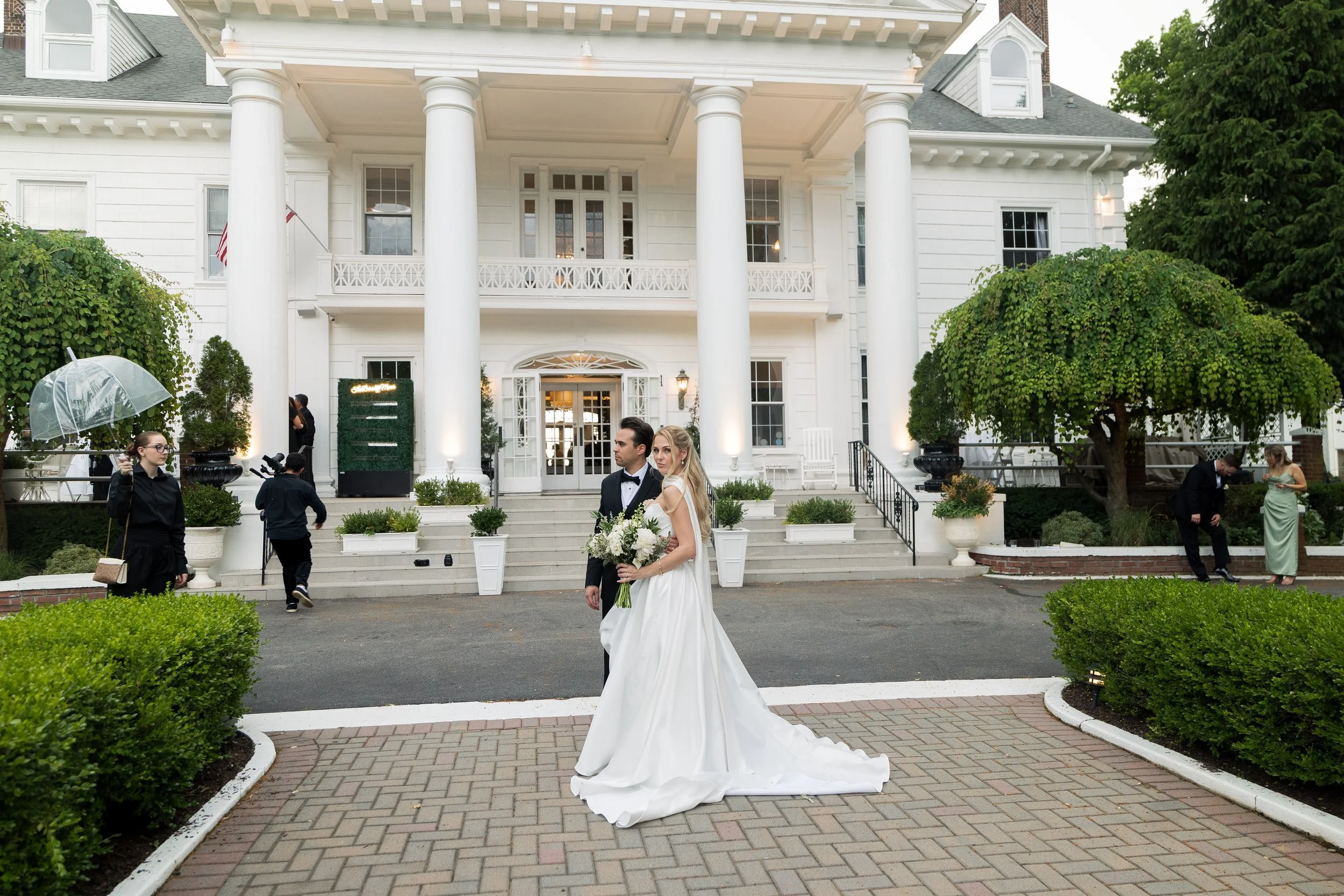 A bride in a white wedding gown holding a bouquet stands with a groom in a tuxedo on the steps of a large white house with columns. Wedding guests and staff are nearby, with some holding umbrellas, in a garden setting.