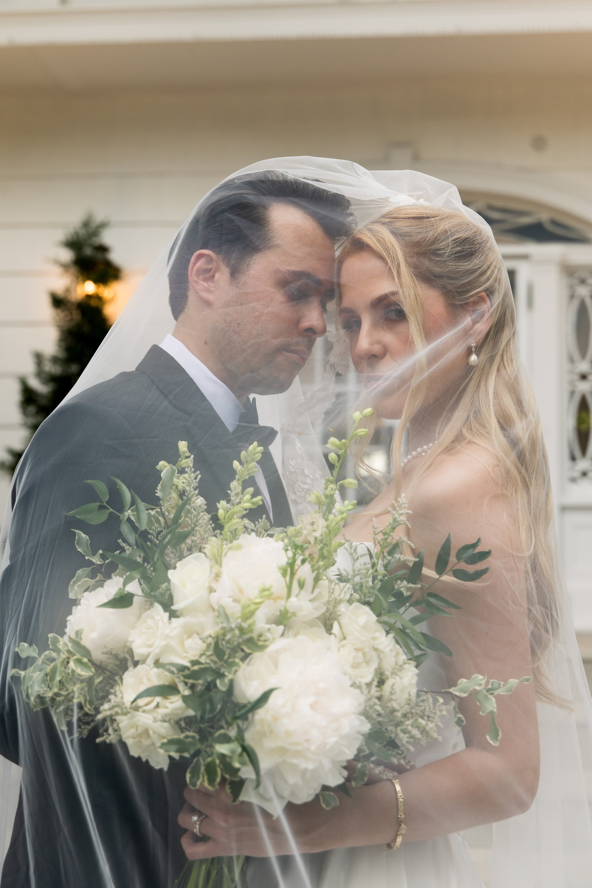 A bride and groom sharing a close moment under a wedding veil. The bride has blonde hair, is wearing pearls, and holding a bouquet of white flowers and greenery. The groom has dark hair and is wearing a black tuxedo and bow tie. They are standing out
