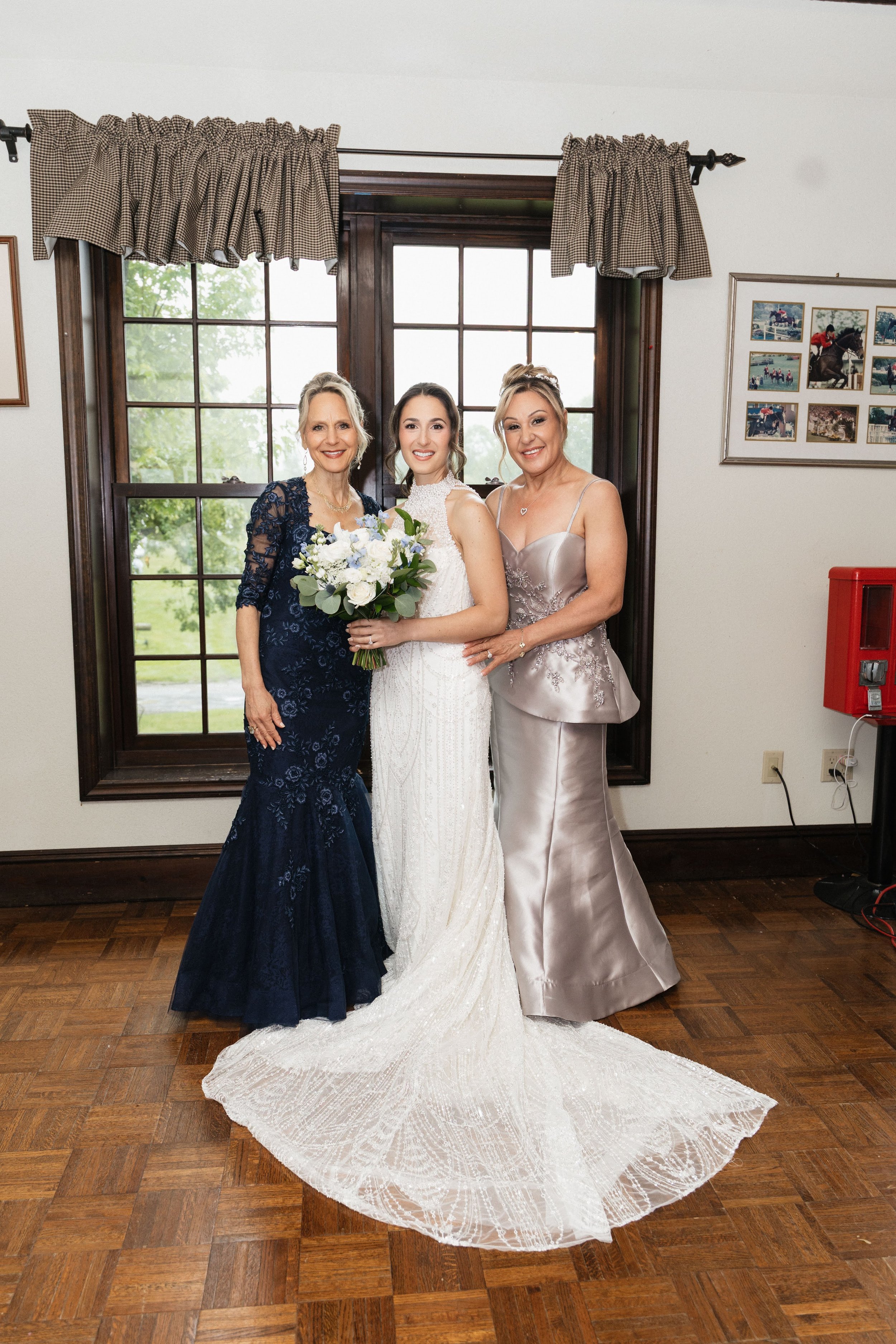 Three women standing together indoors in front of a large window with dark wood framing, each wearing elegant formal dresses. The woman in the middle is holding a bouquet of white flowers and wearing a wedding gown, suggesting a bridal occasion. The 
