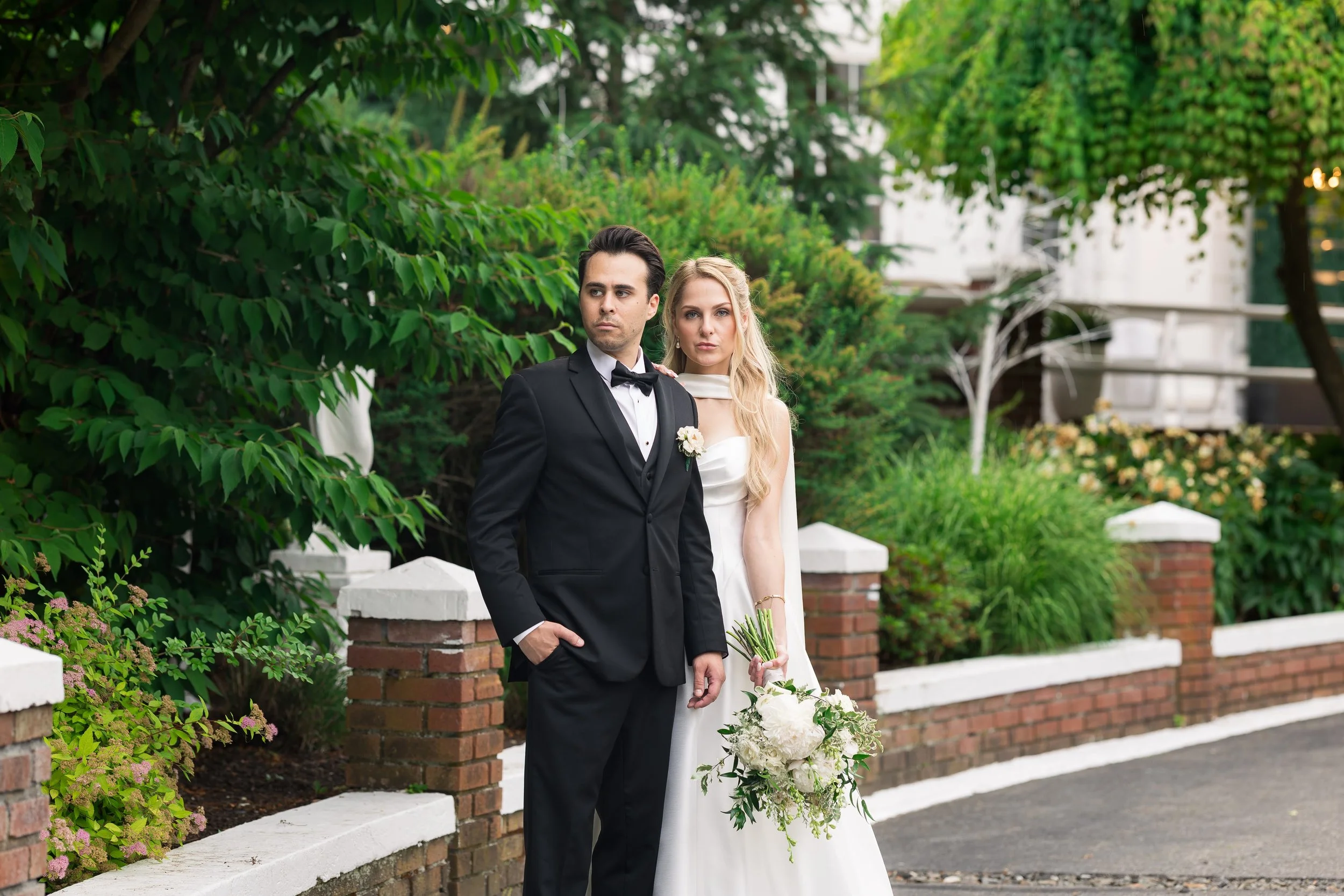 A couple dressed in formal wedding attire standing outdoors in front of green foliage and a brick wall with white trim. The groom is wearing a black tuxedo with a bow tie, and the bride is in a white wedding gown holding a bouquet of white flowers. T