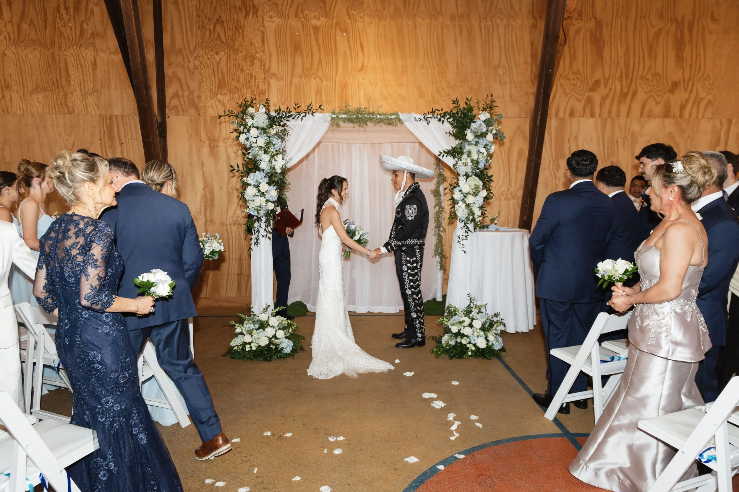 A wedding ceremony with a bride and groom exchanging vows under an arch decorated with white and green flowers, surrounded by guests in formal attire in a wooden indoor venue.