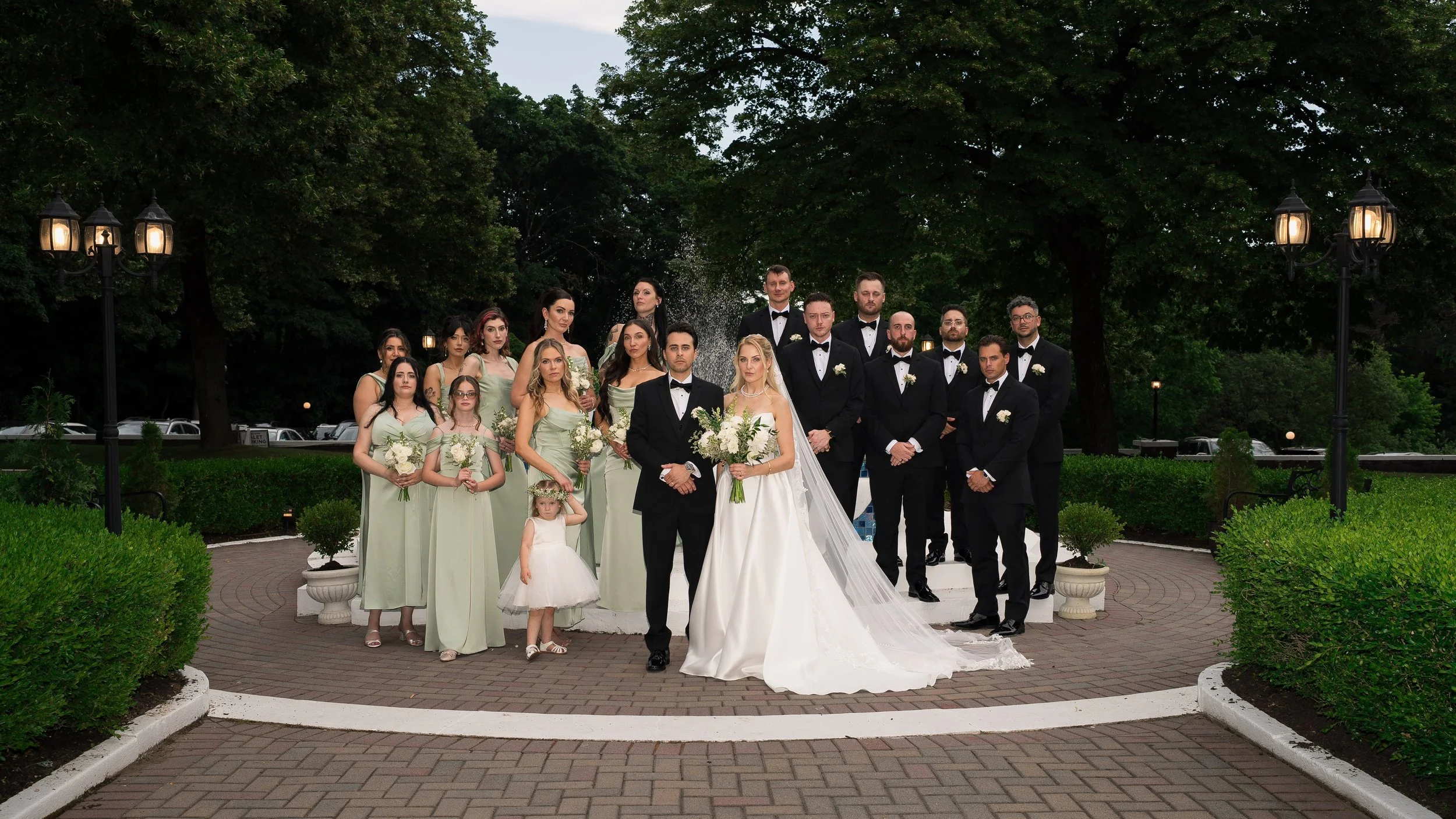 A group of people in wedding attire, including a bride and groom, posing outdoors in a park-like setting with trees and lamps, for a wedding photo.