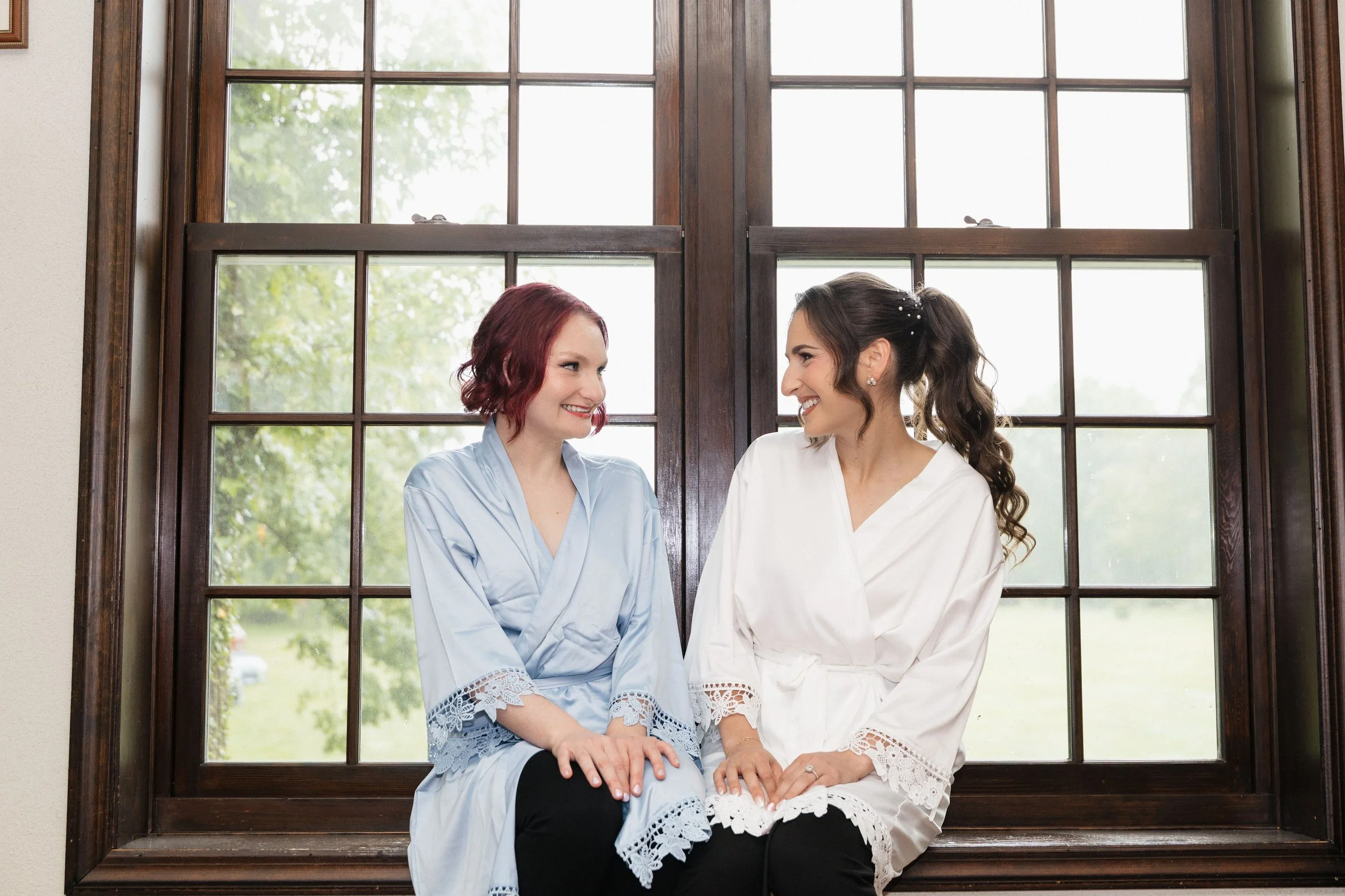 Two women sitting together by a large window, smiling at each other. One woman has red hair and is wearing a light blue dress, while the other has brown hair tied in a ponytail and is wearing a white dress with lace details.