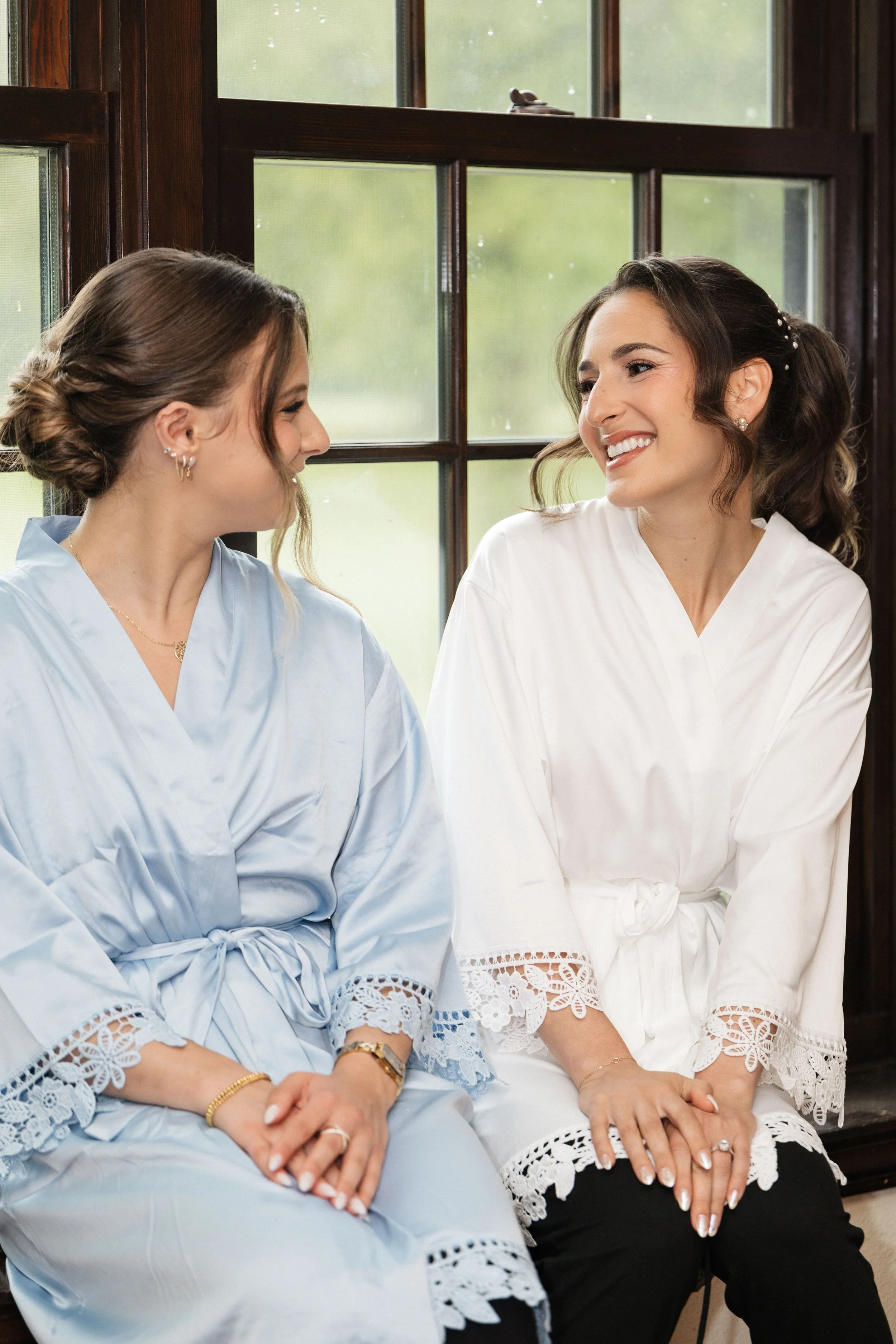 Two women sitting side by side in front of a window, looking at each other with smiles. One is dressed in light blue and the other in white with lace details.