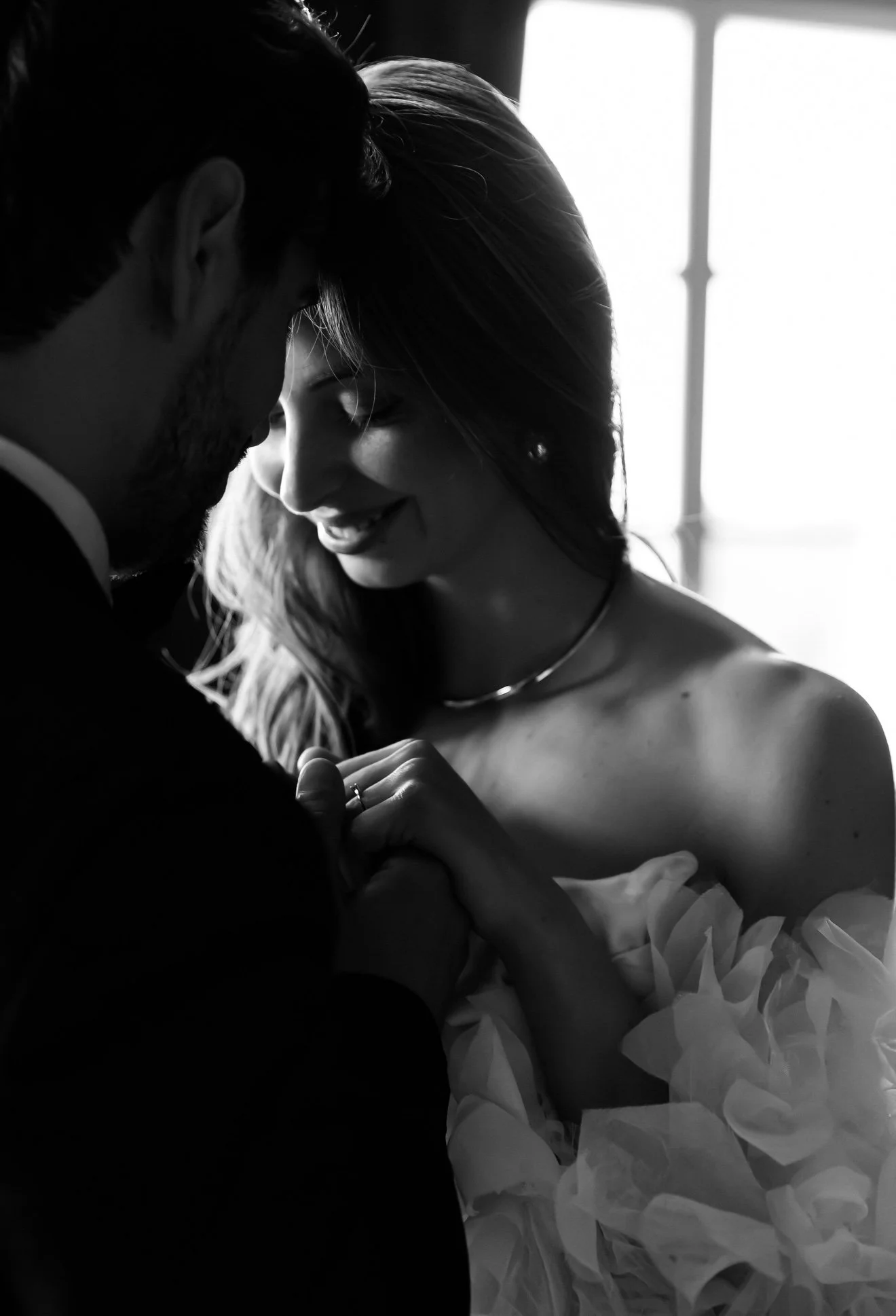 A couple sharing an intimate moment, with their foreheads and noses touching, smiling softly, in a dimly lit room with a window in the background.